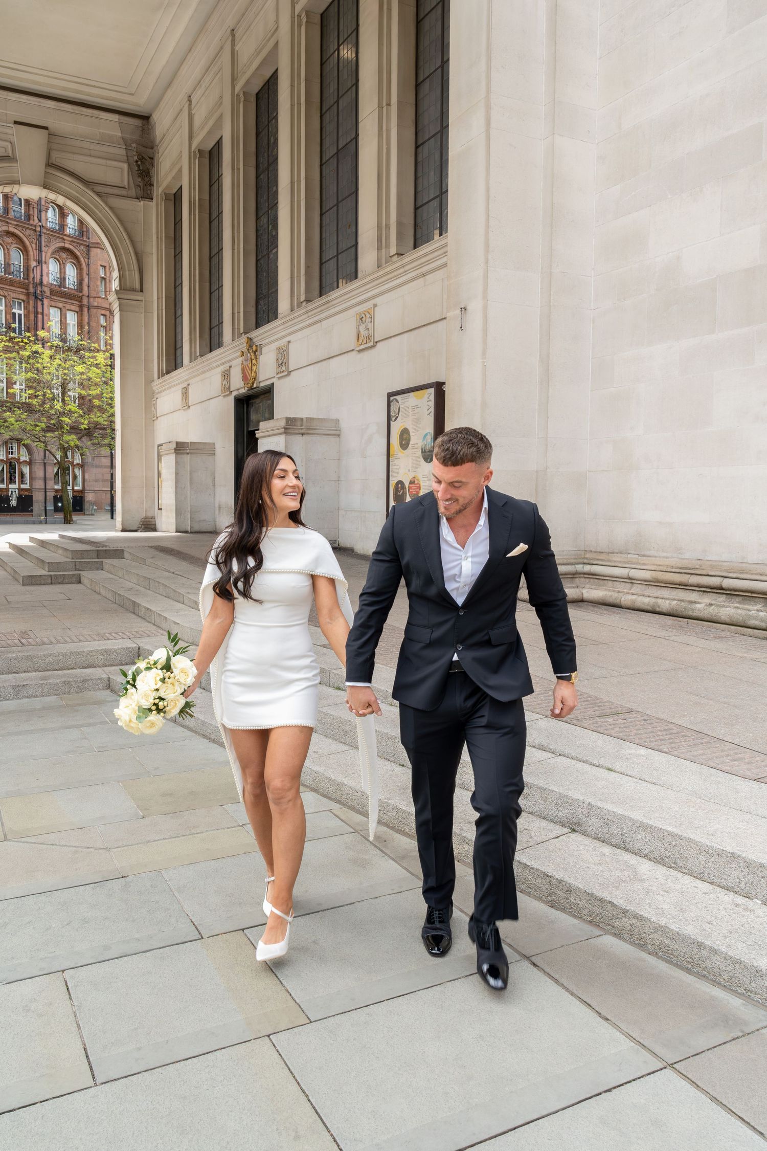 Bride in white mini dress and groom in navy suit walk hand in hand outside a grand stone building.