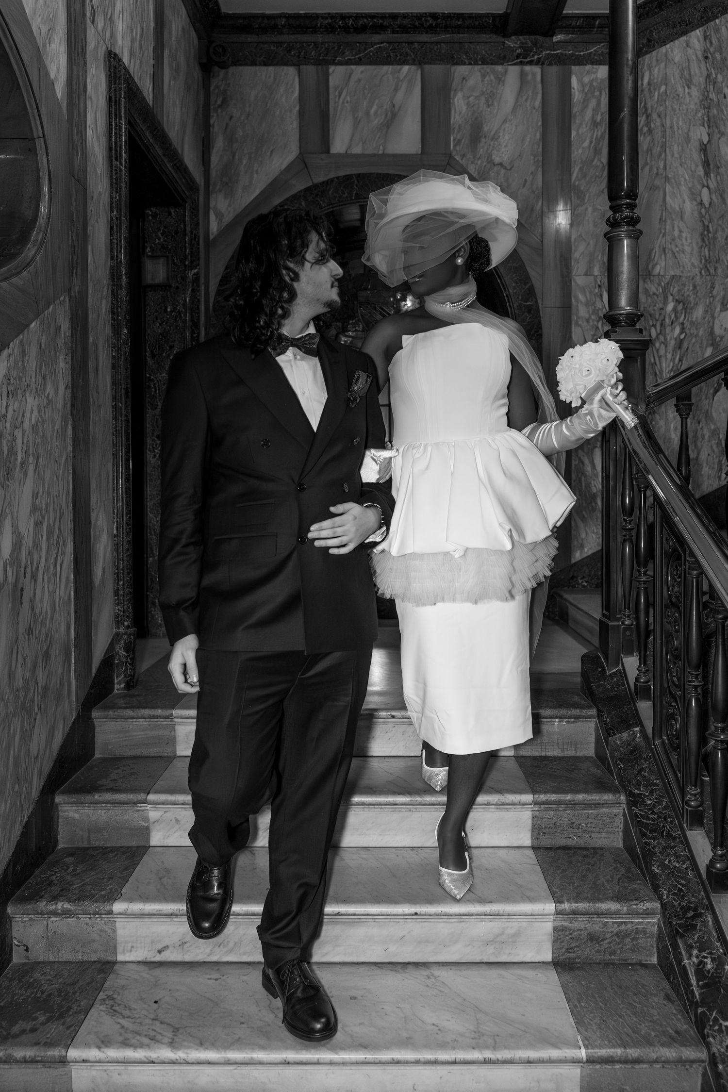 Black and white photo of a bride and groom walking up marble stairs, bride holding bouquet wearing short dress and veil.