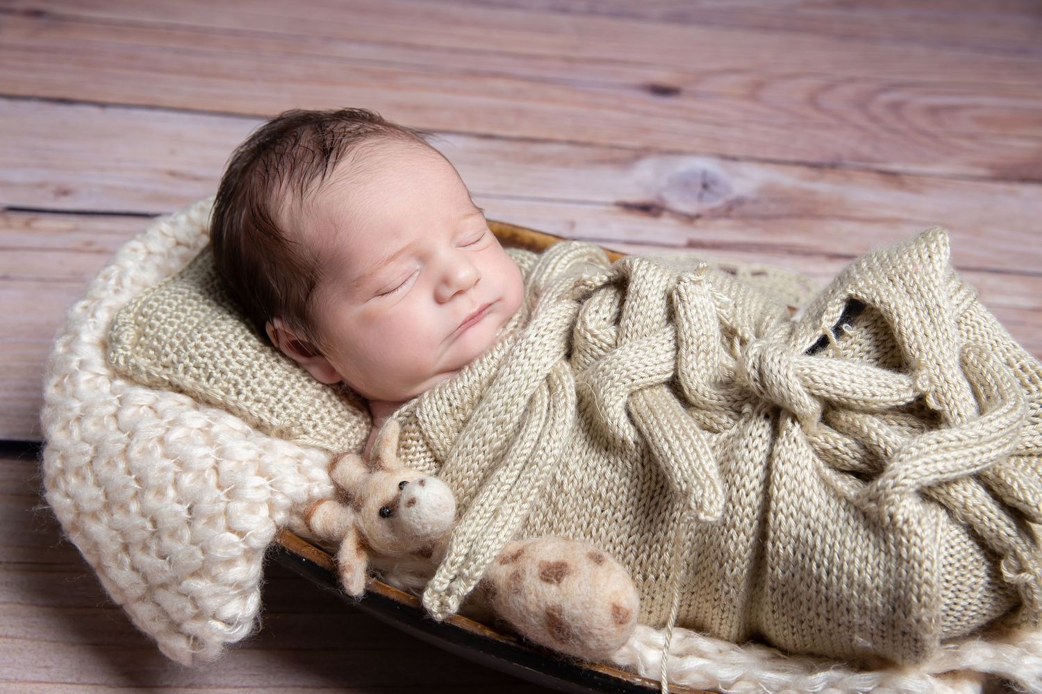 A sleeping newborn baby wrapped in cozy cream knit blankets rests peacefully on a wooden floor.