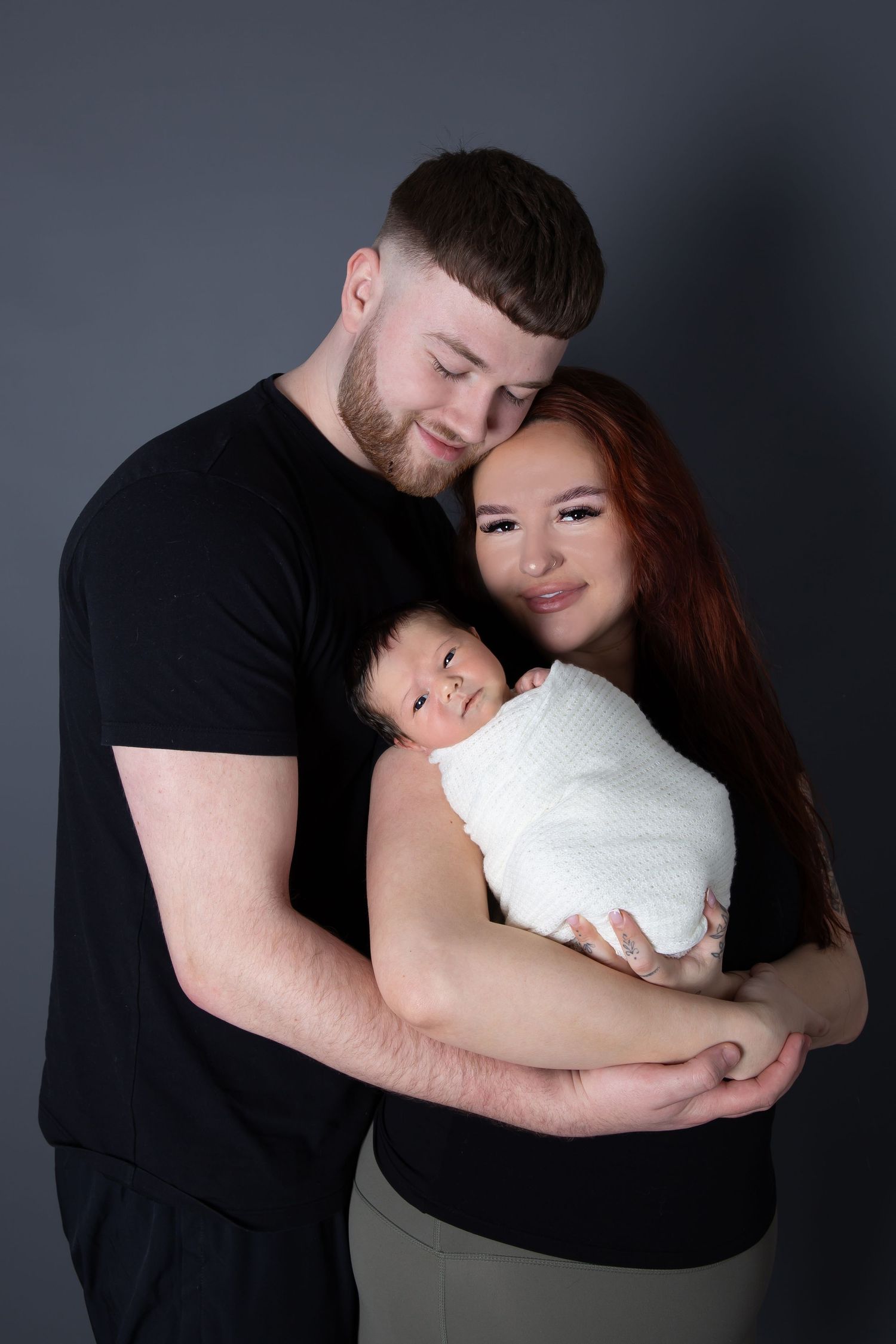 A couple embraces while holding their swaddled newborn baby against a dark background in a studio portrait.