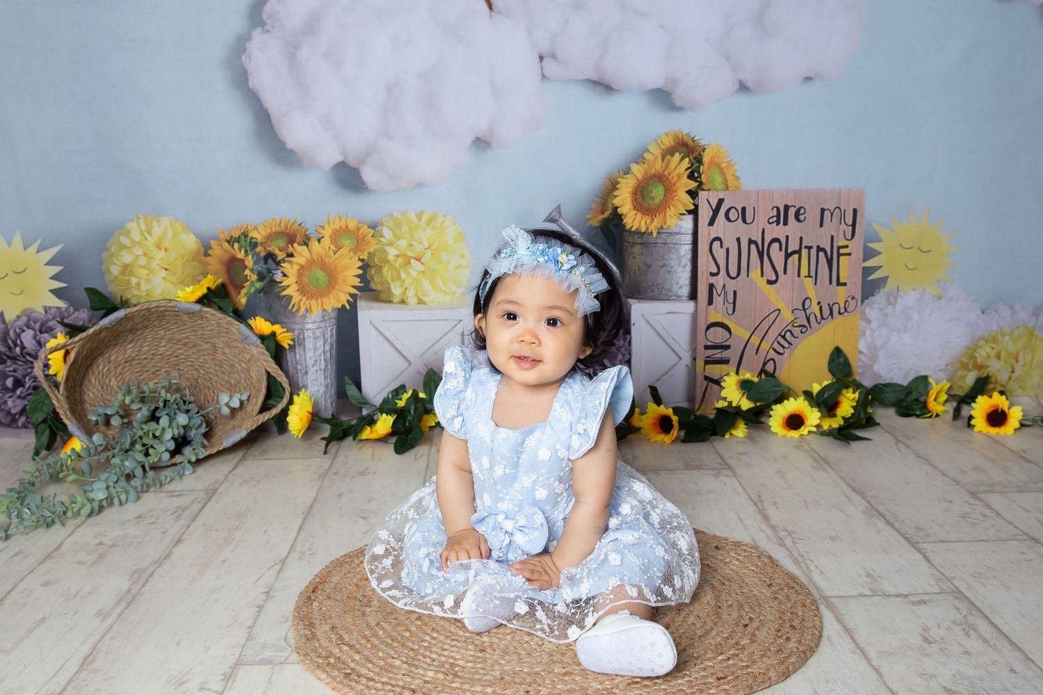 A young child in a blue dress sits on a round rug with sunflower decorations and 'You are my sunshine' sign in the background.