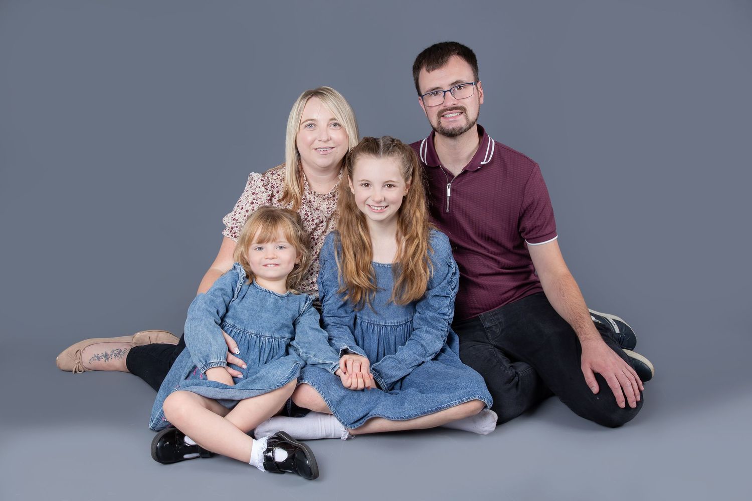 A happy family sits together wearing coordinated blue and burgundy outfits against a gray studio background.