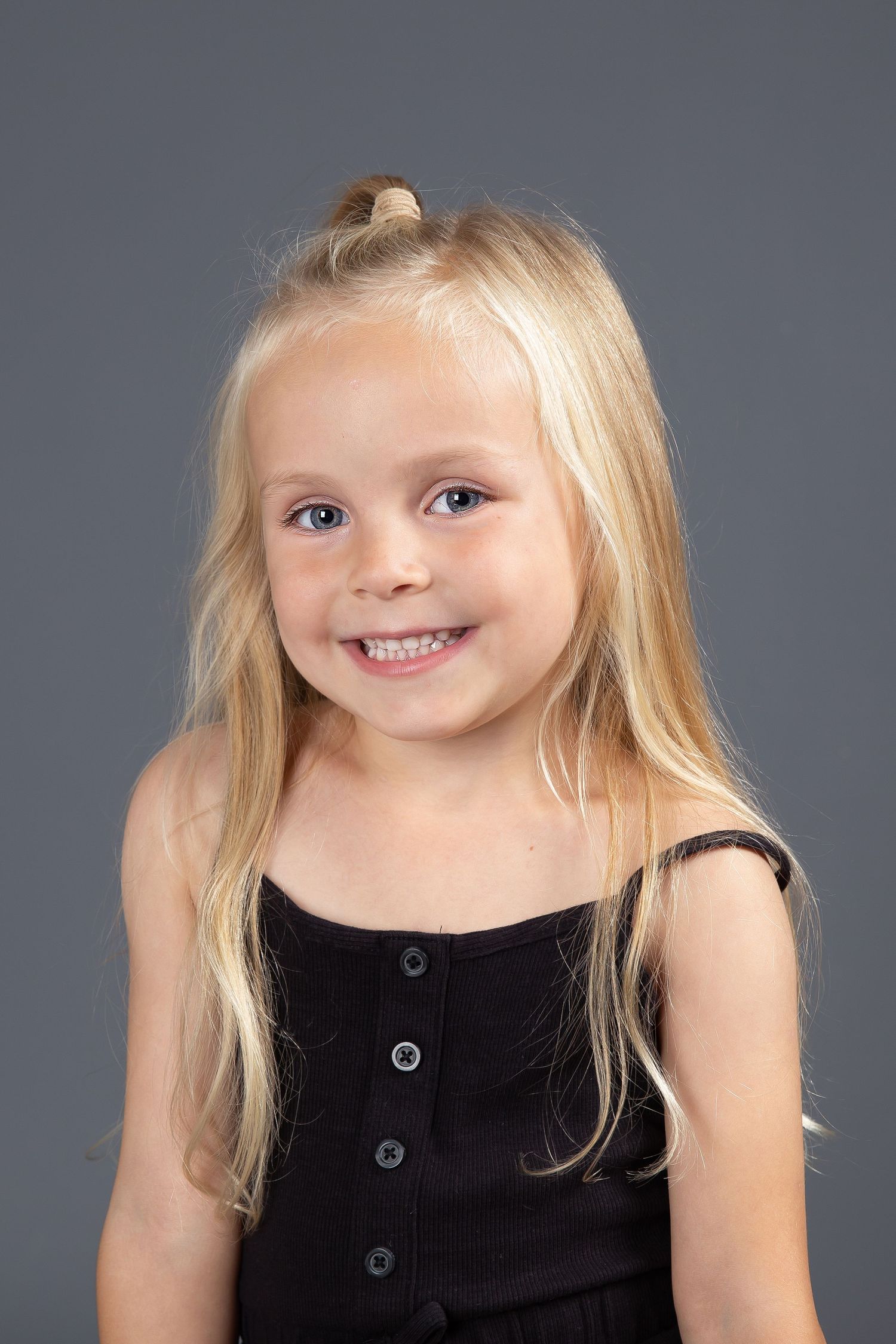 A young child with long blonde hair wearing a black button-up dress smiles at the camera against a gray background.