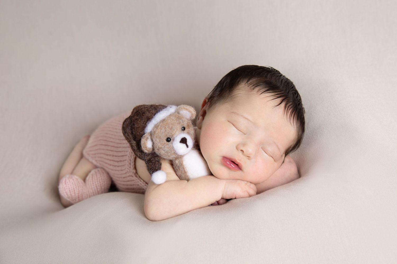 A sleeping newborn wearing a pink knit outfit cuddles with a small teddy bear on a cream background.