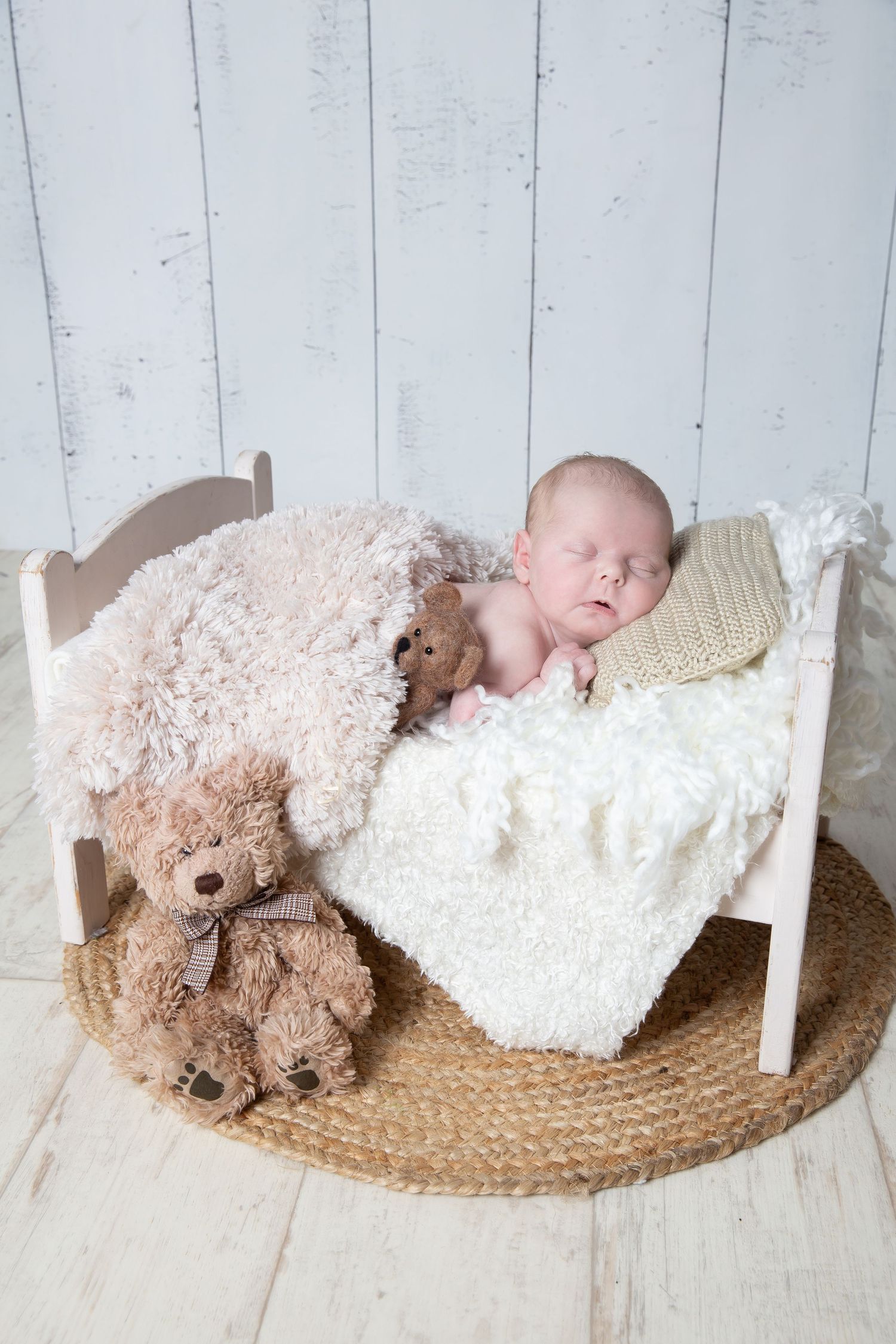 Sleeping baby in white bed with teddy bears and plush toys on woven mat against rustic wooden wall background.
