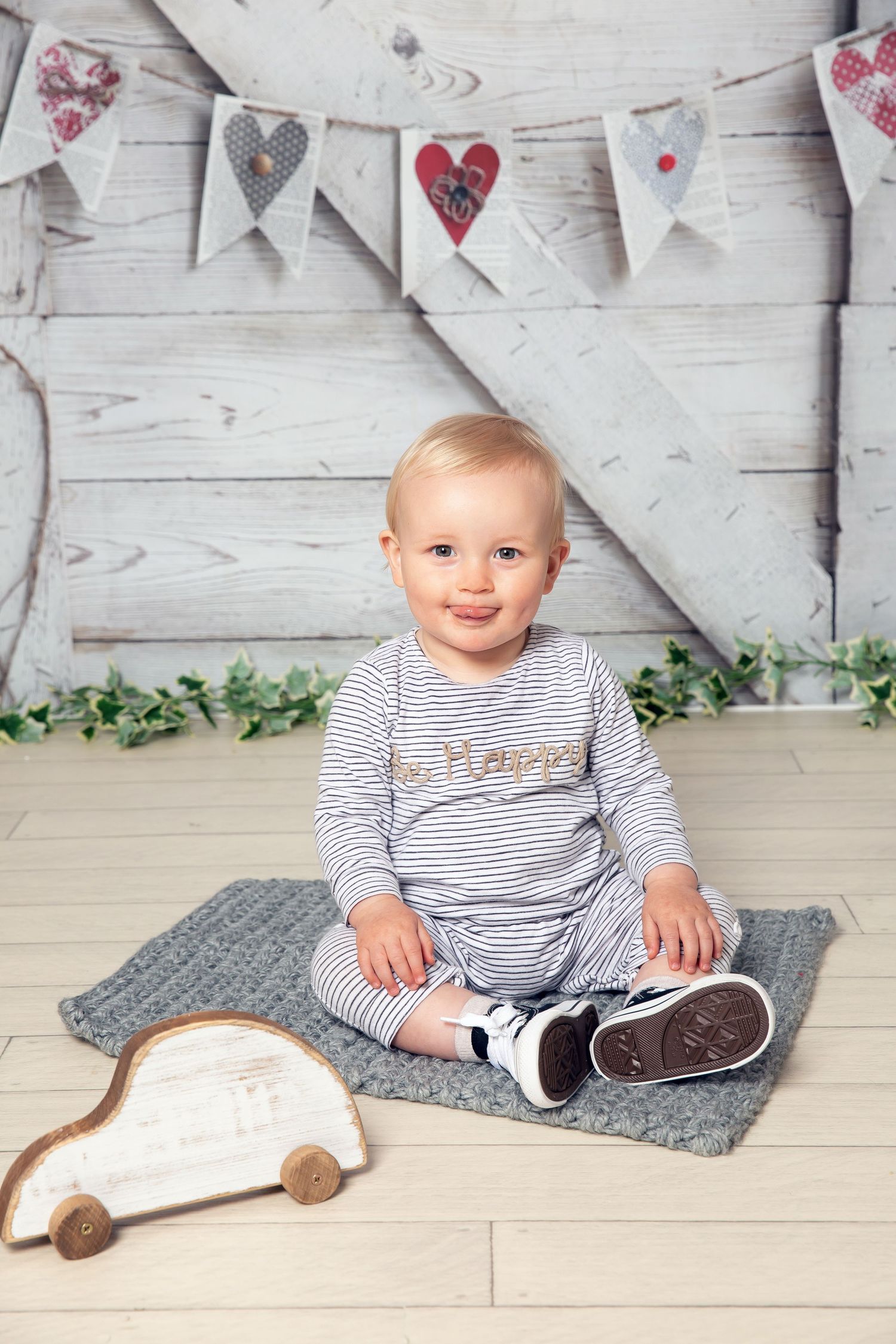 Toddler in striped outfit sits on gray blanket under heart banner decorations with wooden toy car nearby.