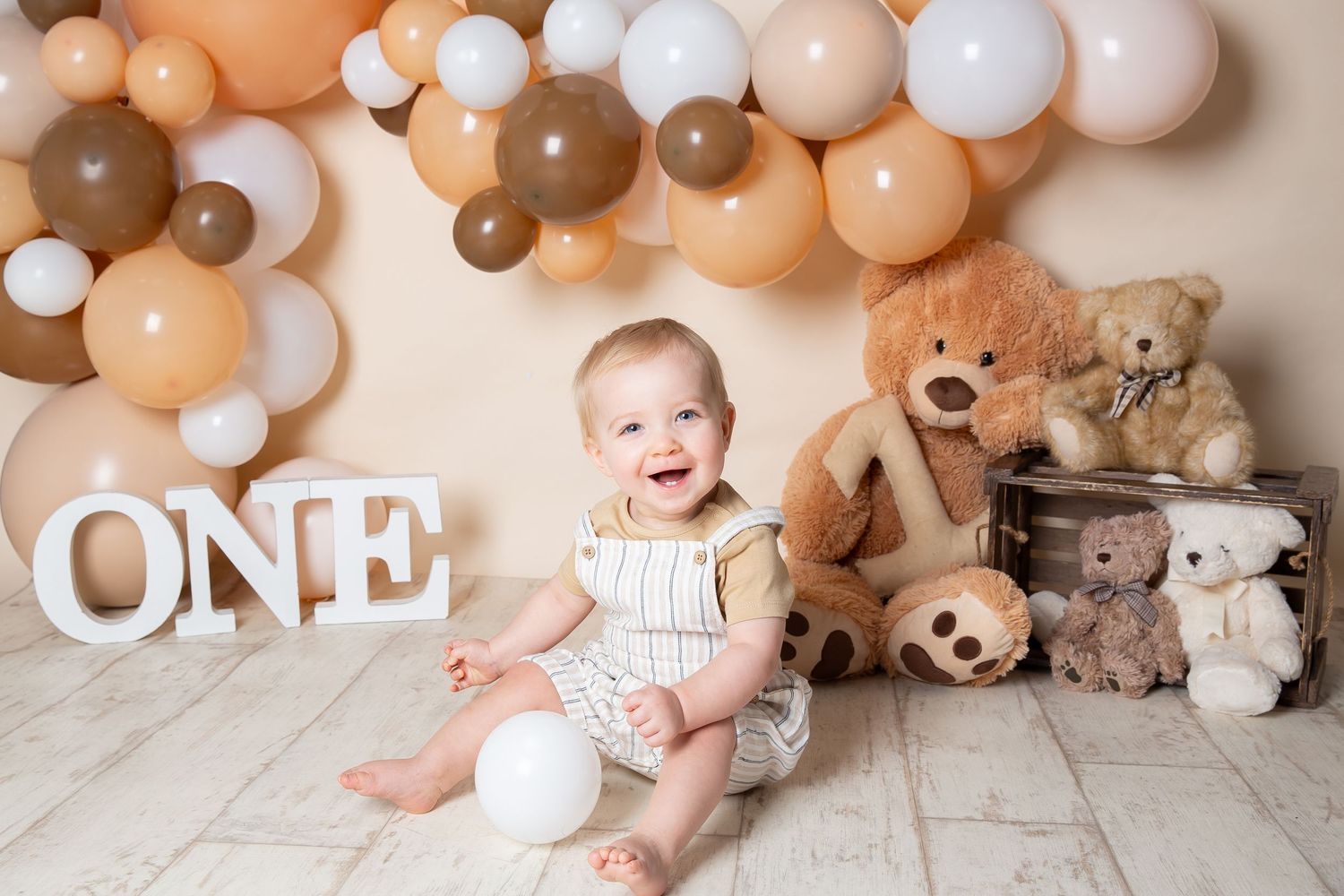 A birthday photo setup with white letters spelling ONE, neutral balloon arch, and stuffed teddy bears against a beige backdrop.
