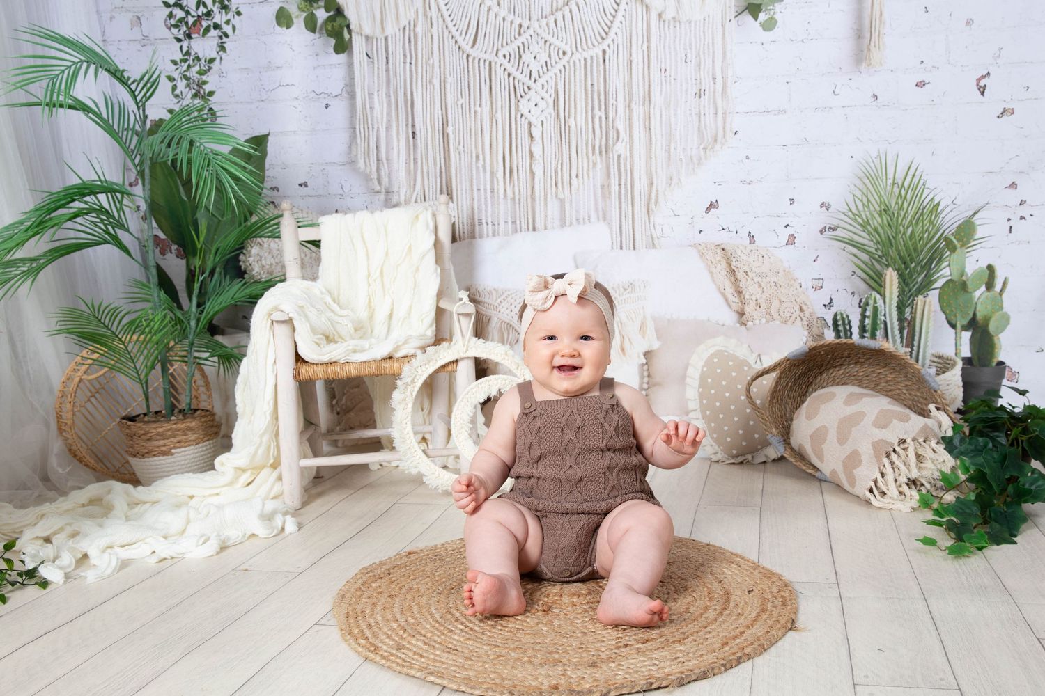 Adorable baby in brown outfit lies in minimalist white nursery setting surrounded by green plants and natural decor.