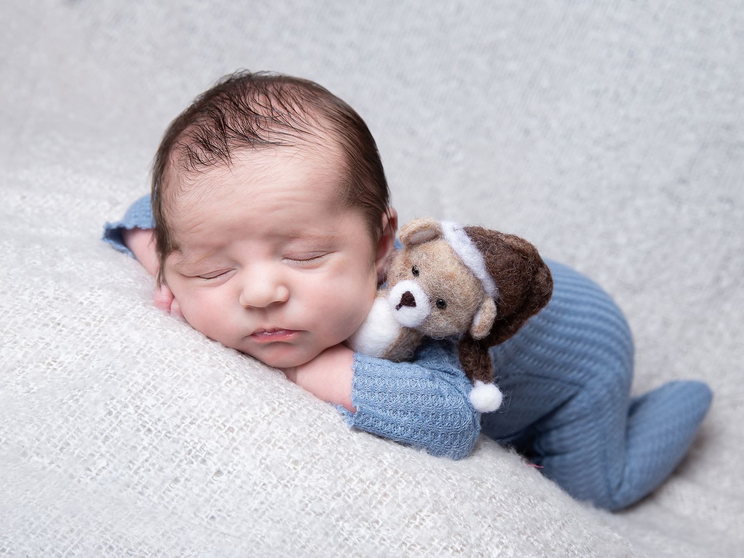 Sleeping newborn baby wearing a blue knit sweater rests peacefully on white textured fabric.