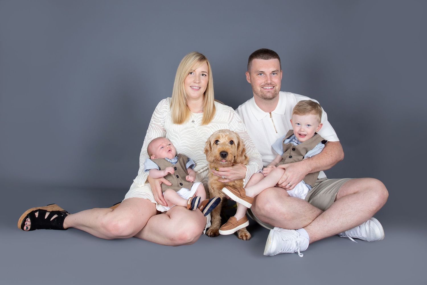 A family in white clothing poses together with their golden retriever puppy on a gray backdrop for a photo series.