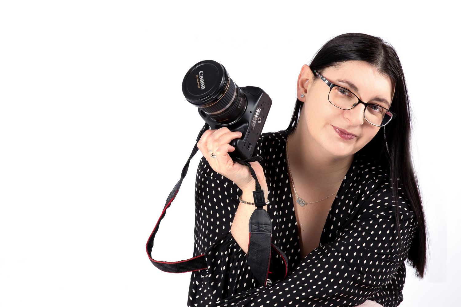 Professional photographer wearing polka dot dress holds DSLR camera while posing against white background.