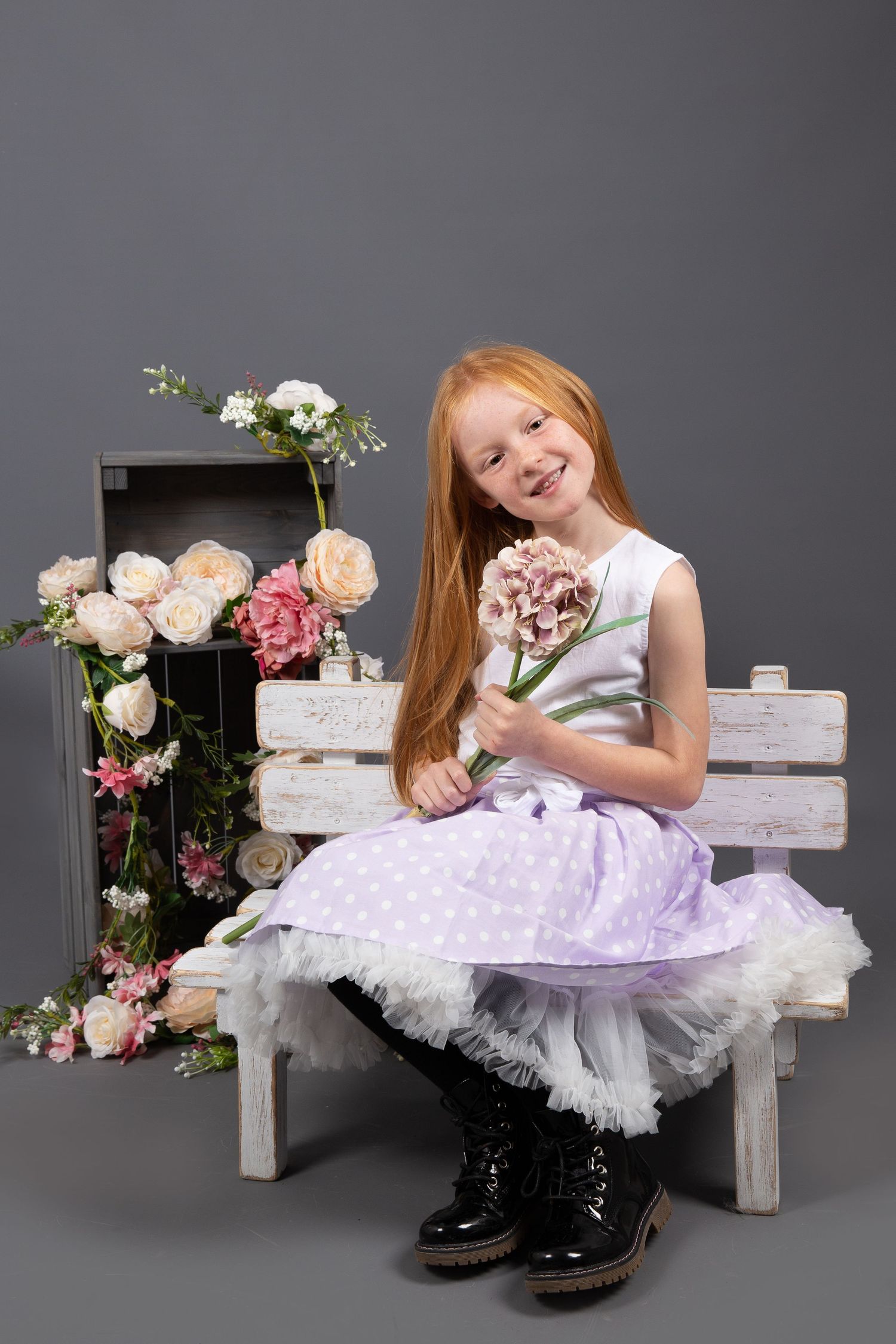 A rustic wooden bench decorated with pink roses and flowers creates a whimsical springtime scene against a gray backdrop.