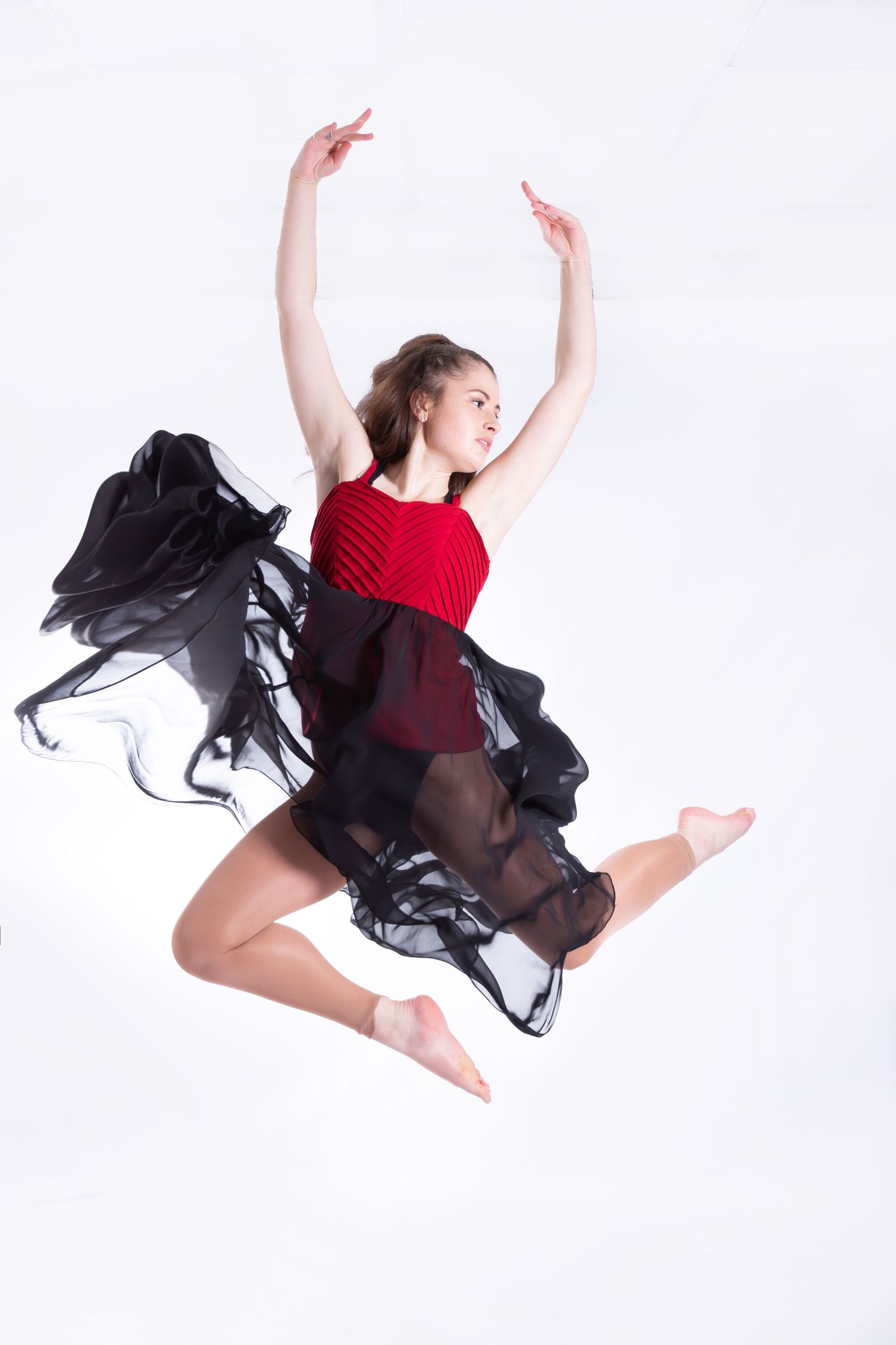 Dancer in red leotard and flowing black sheer skirt performs graceful leap against white background.