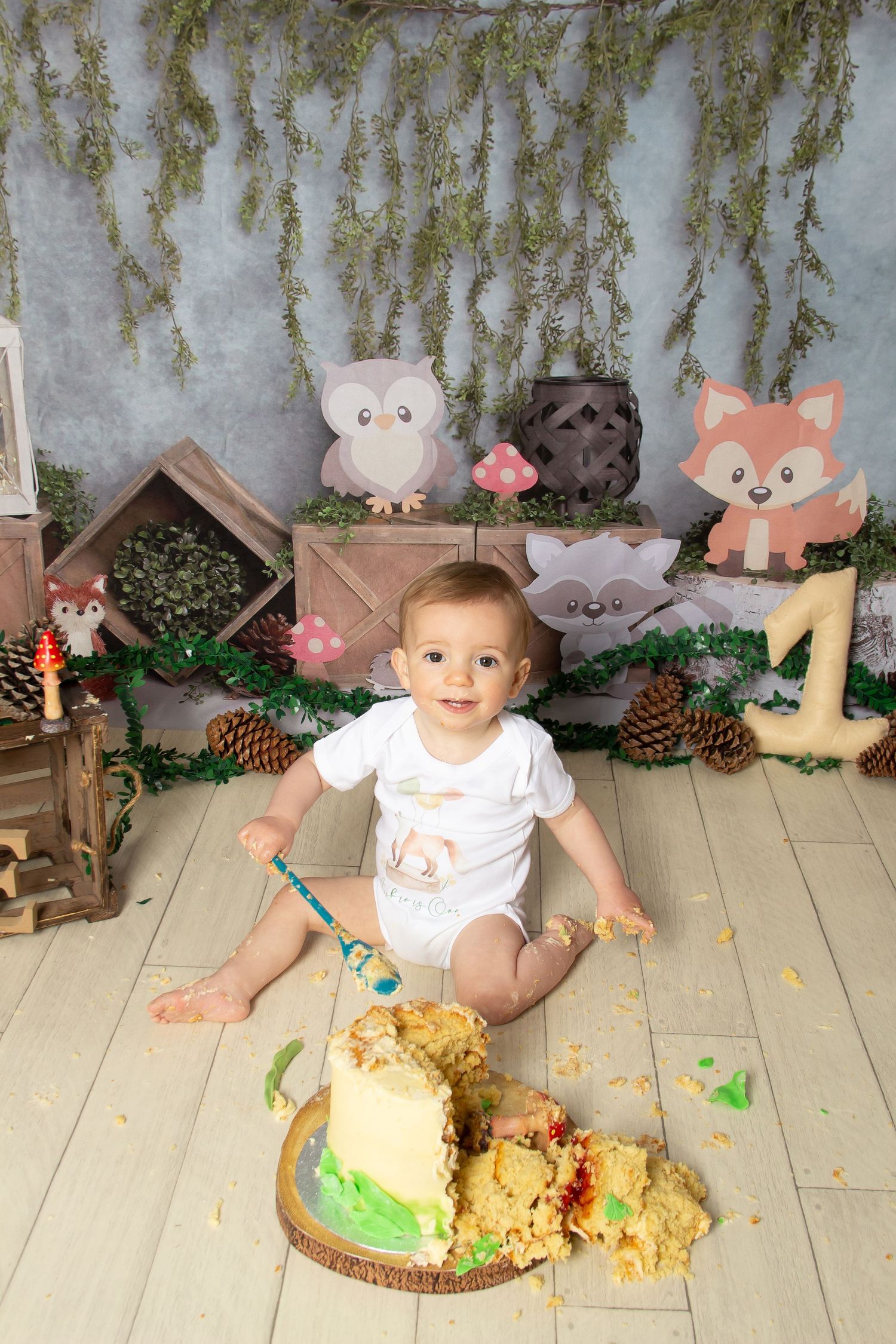 Woodland themed cake smash photo session setup with forest animals, pinecones and greenery on light wooden flooring.