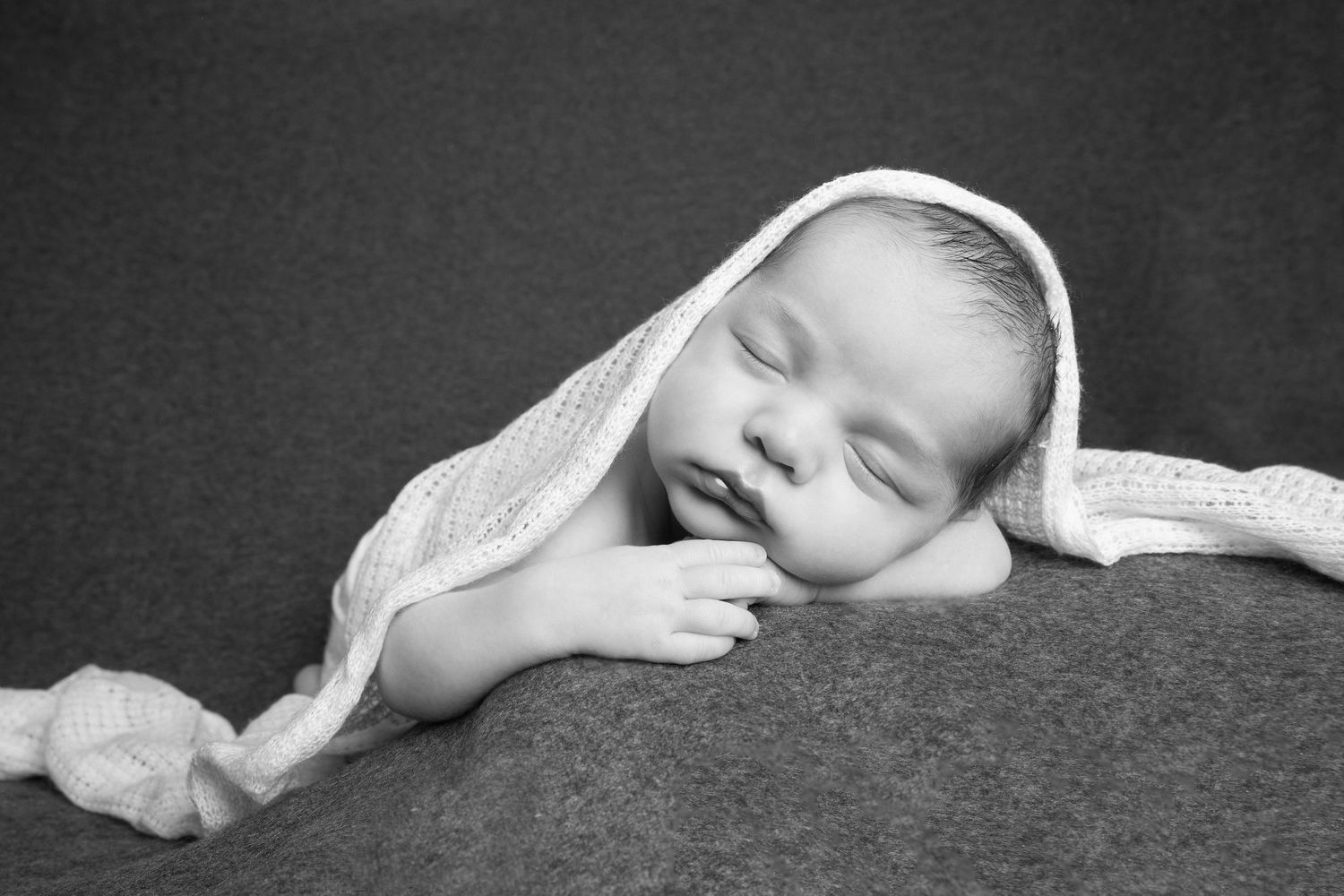 A tranquil black and white artistic portrait of a sleeping baby draped in soft white fabric lying against a dark backdrop.