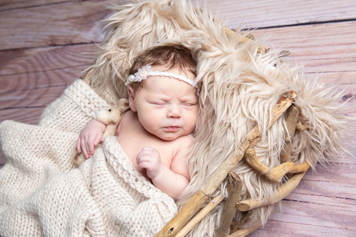 A sleeping baby wearing a knitted headband lies on a wooden floor next to small plush toy animals.