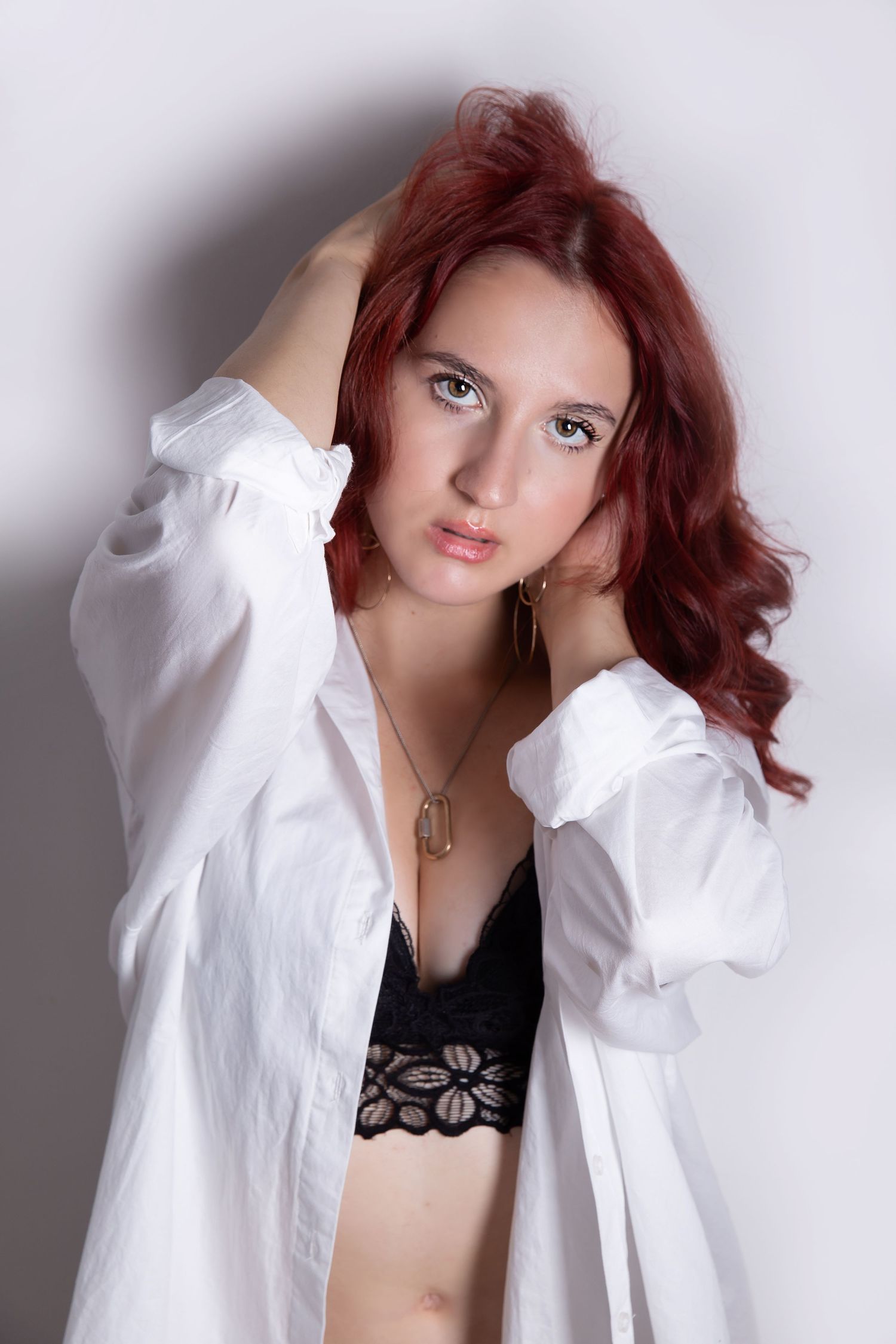 Portrait of a person with red hair wearing a white button-down shirt and black lace top against a light backdrop.
