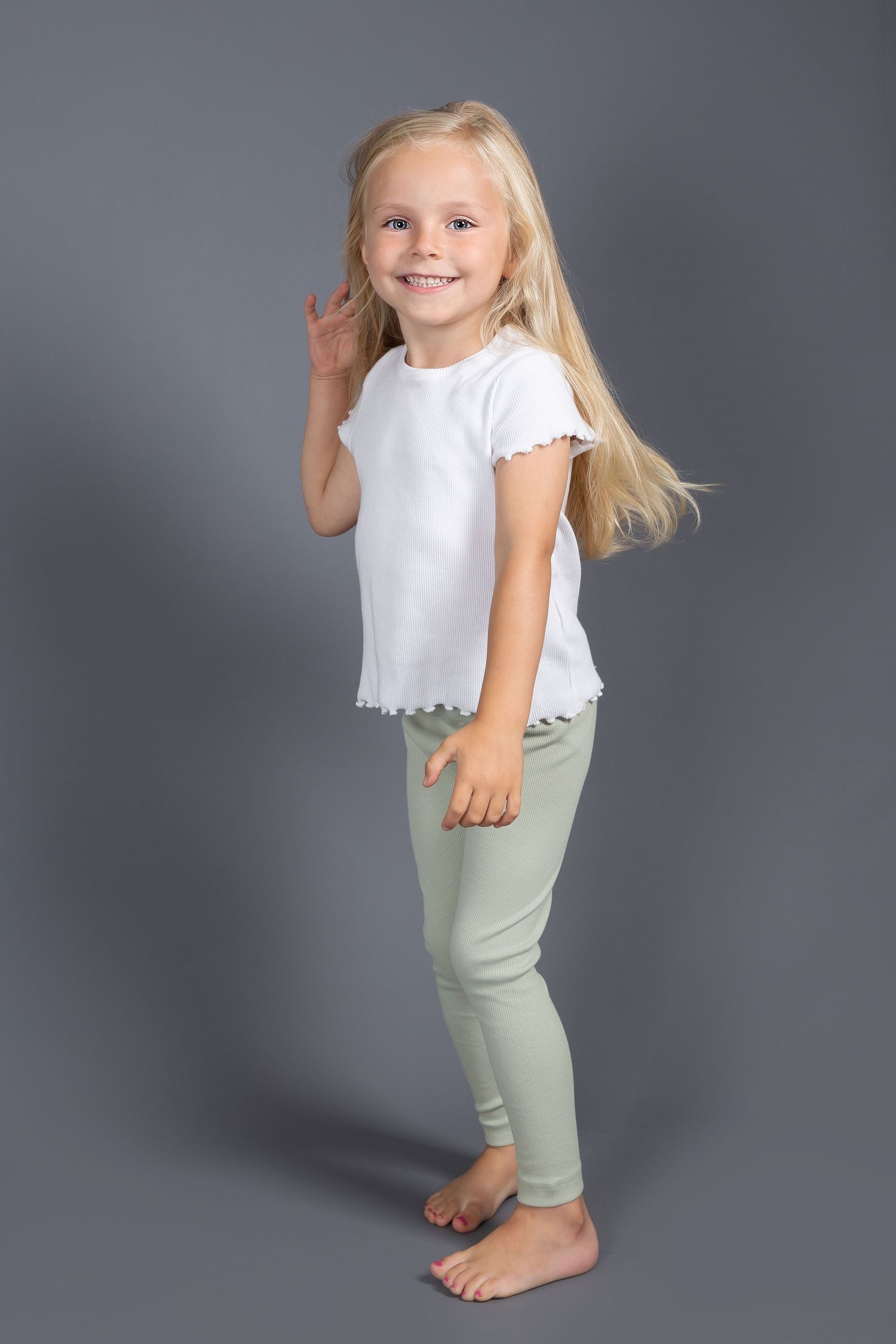 A young child wearing a white eyelet top and sage green pants poses playfully in front of a gray background.