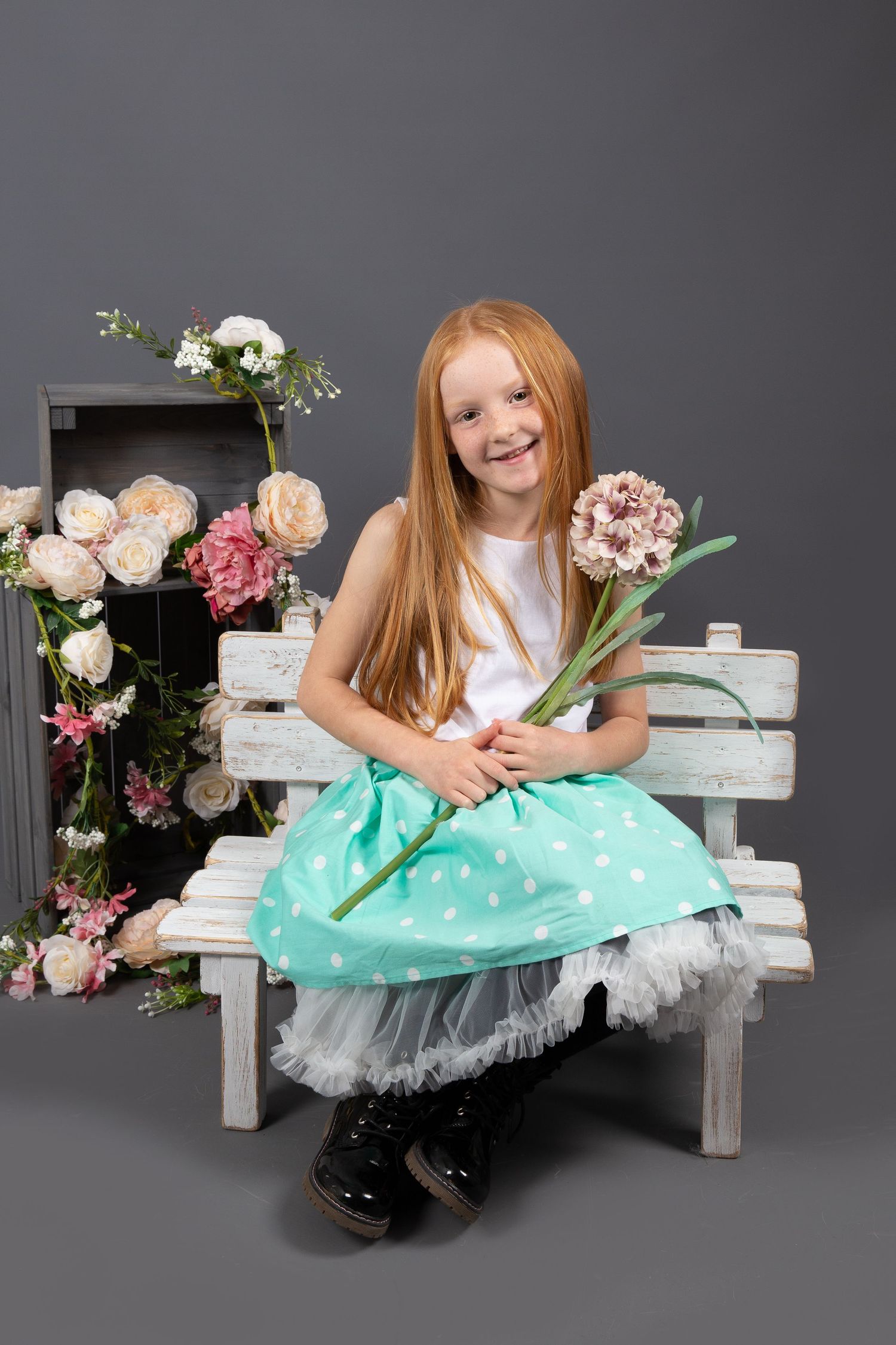 A young person in a mint polka dot skirt and white top sits on a rustic bench surrounded by pink and white flowers.
