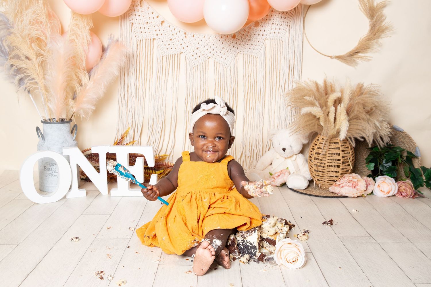 A child in a yellow dress sits on the floor with decorative items and a macrame backdrop for a first birthday celebration.