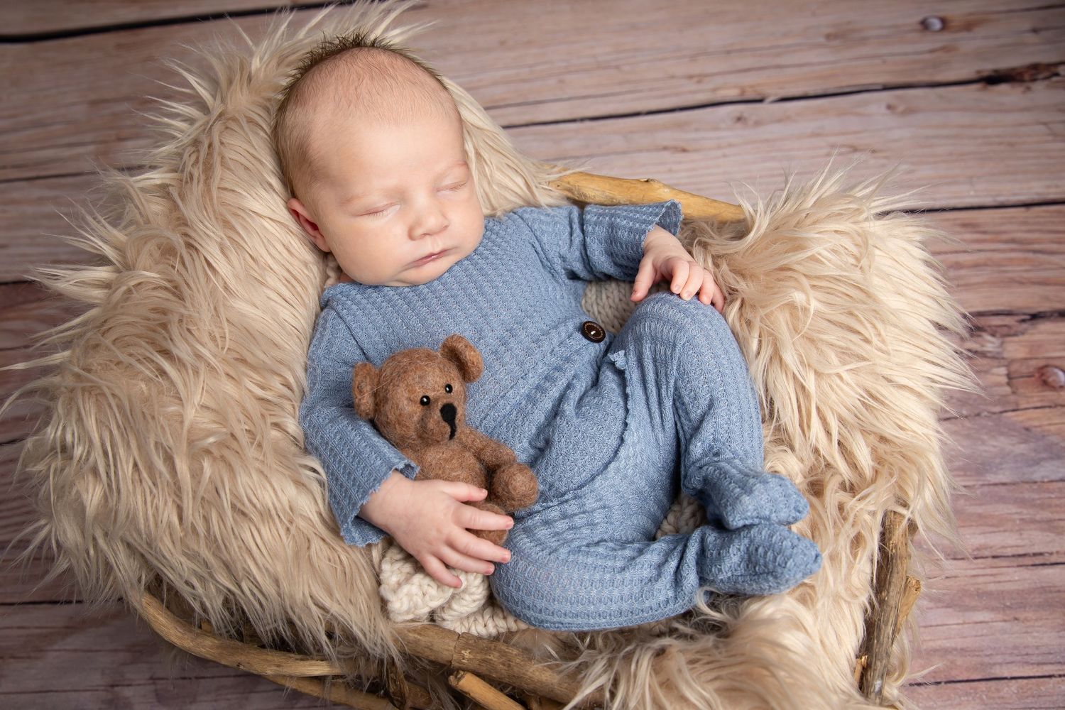 Sleeping baby in blue knit outfit cuddled with teddy bear on furry cushion against wooden backdrop.