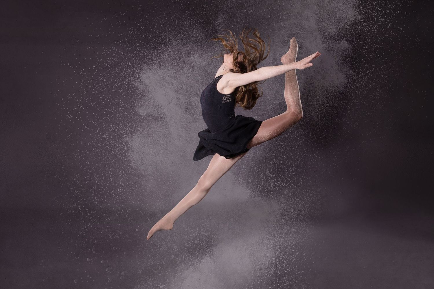 A dancer in a black dress leaps gracefully through a cloud of powder against a dark background.