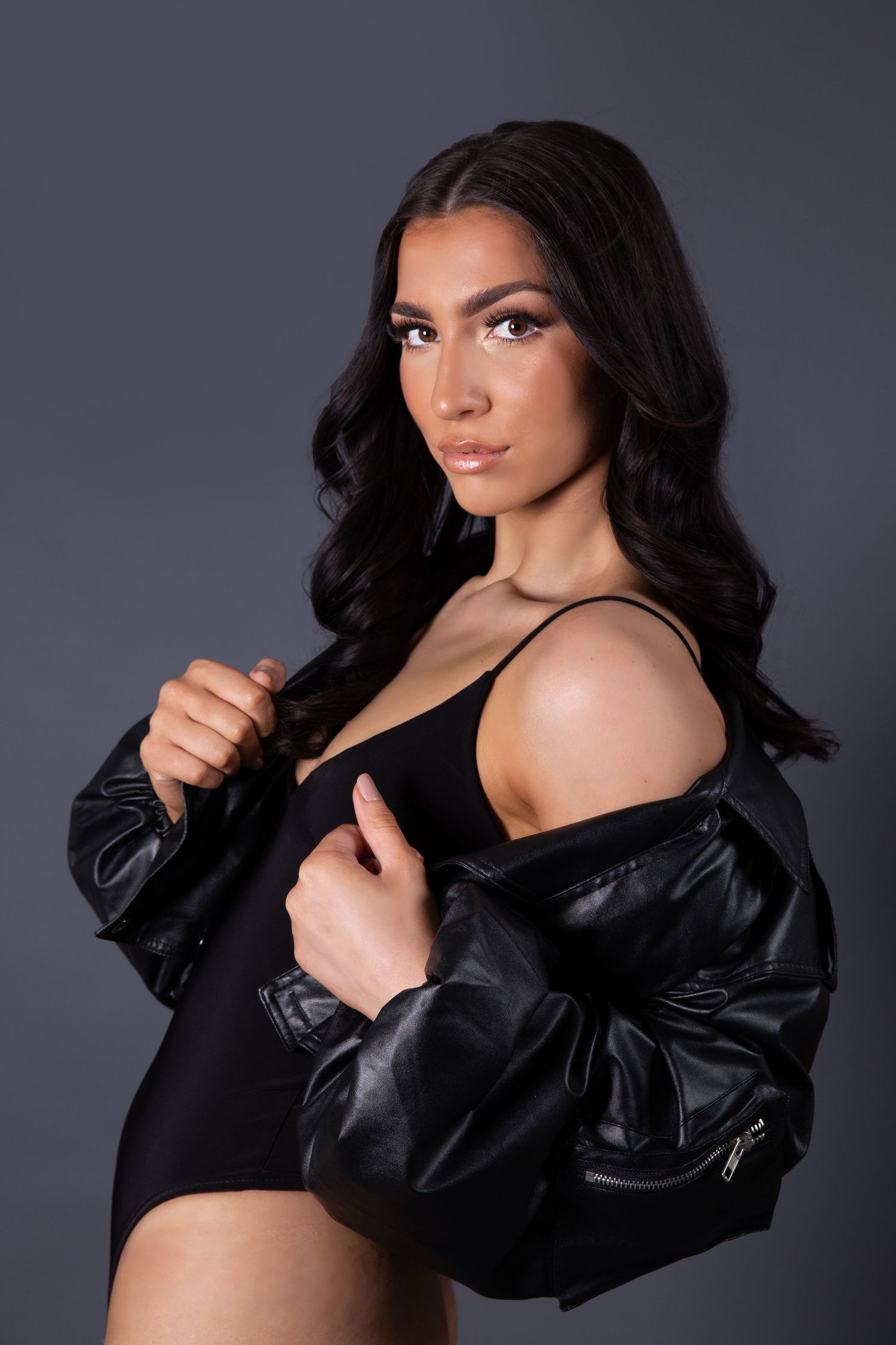 Fashion model poses in black bodysuit and leather jacket against dark studio background.