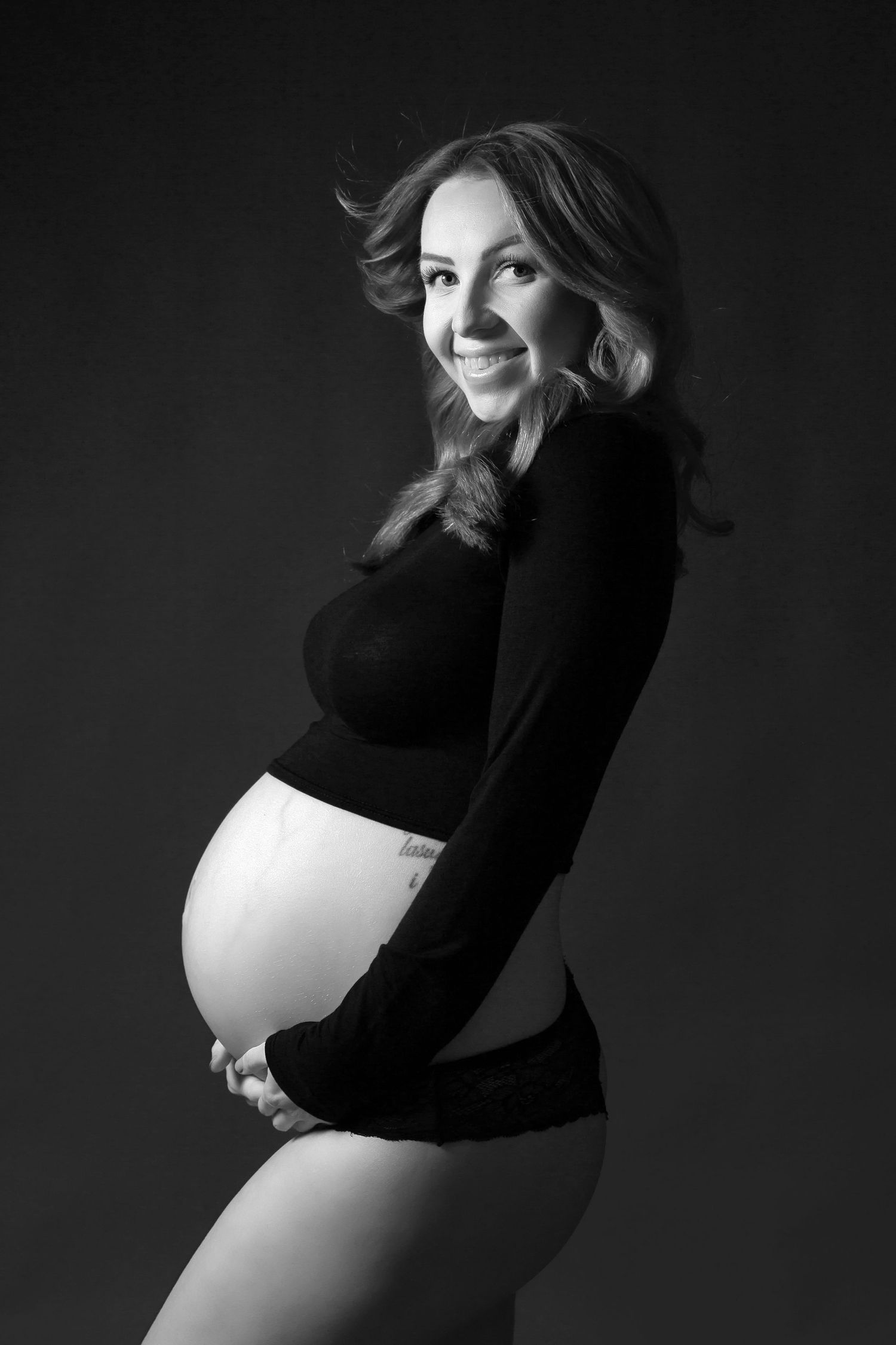 Black and white maternity portrait of smiling woman in dark sweater and underwear against dark background.