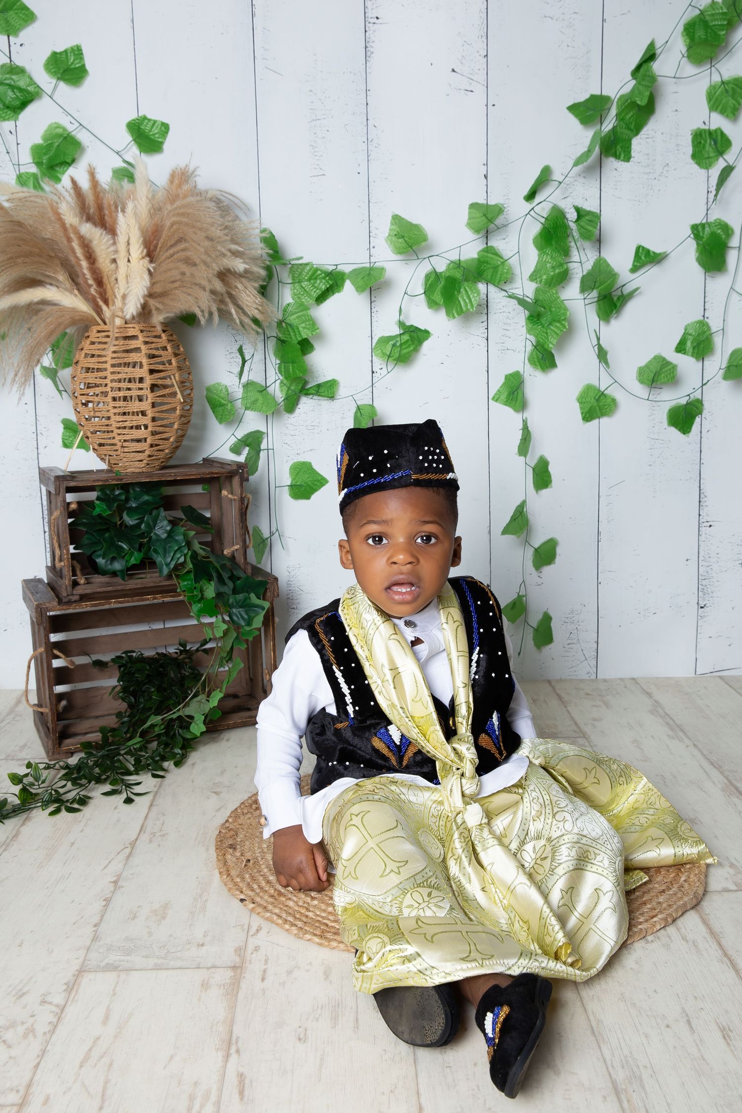 A baby in traditional West African attire with a gold and black embroidered garment sits in front of a rustic backdrop.