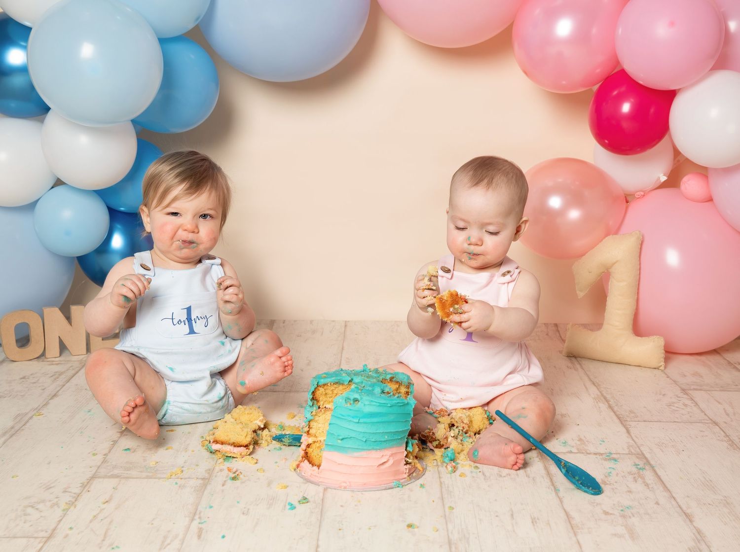 Baby in white outfit sits on floor with cake and colorful balloons during first birthday cake smash photo session.