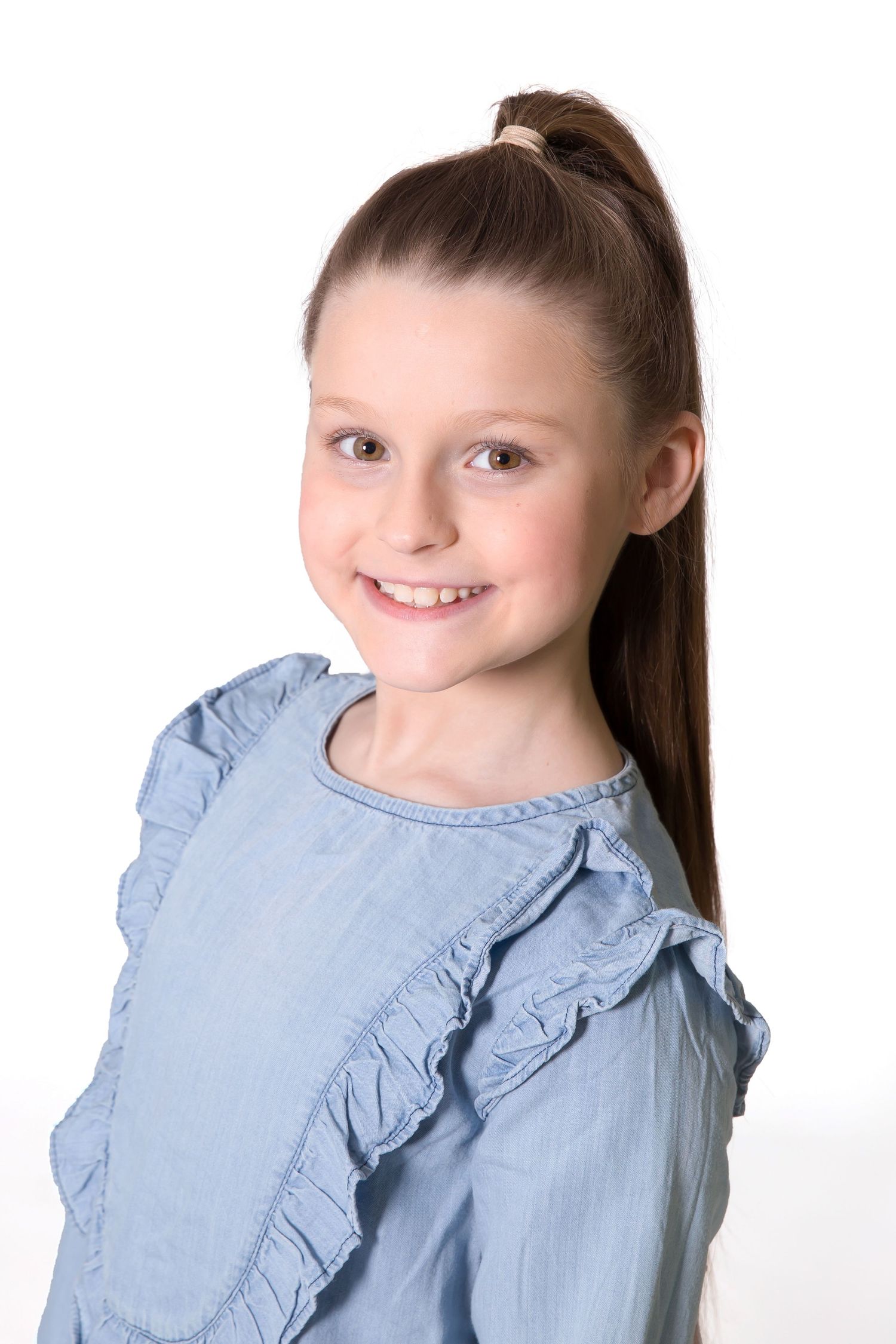 Smiling young girl with brown hair in ponytail wearing light blue ruffled top against white background.