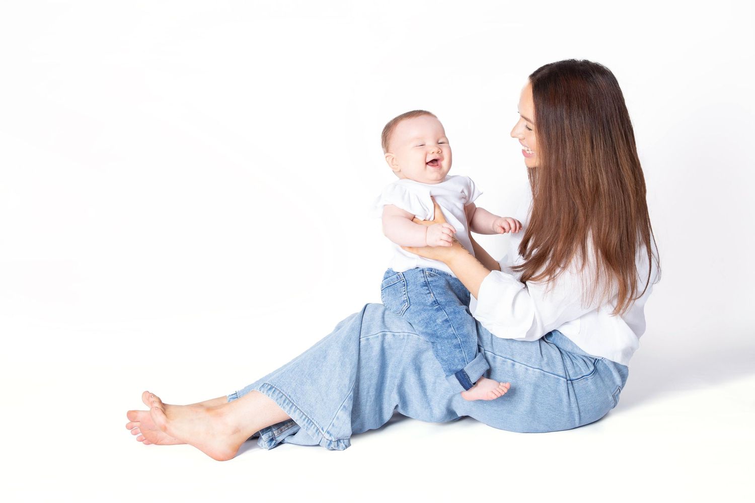 A joyful mother and baby share a playful moment together while sitting on a white floor.