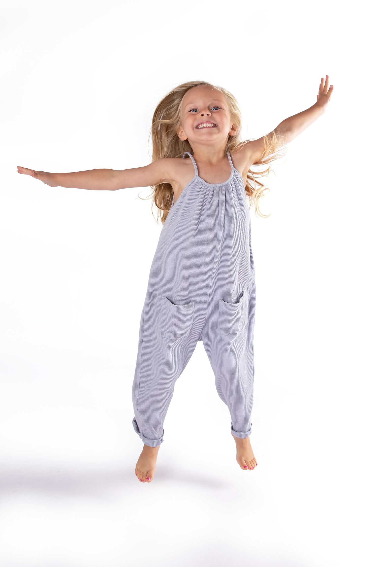 Child jumping joyfully in gray jumpsuit romper against white background with arms outstretched.