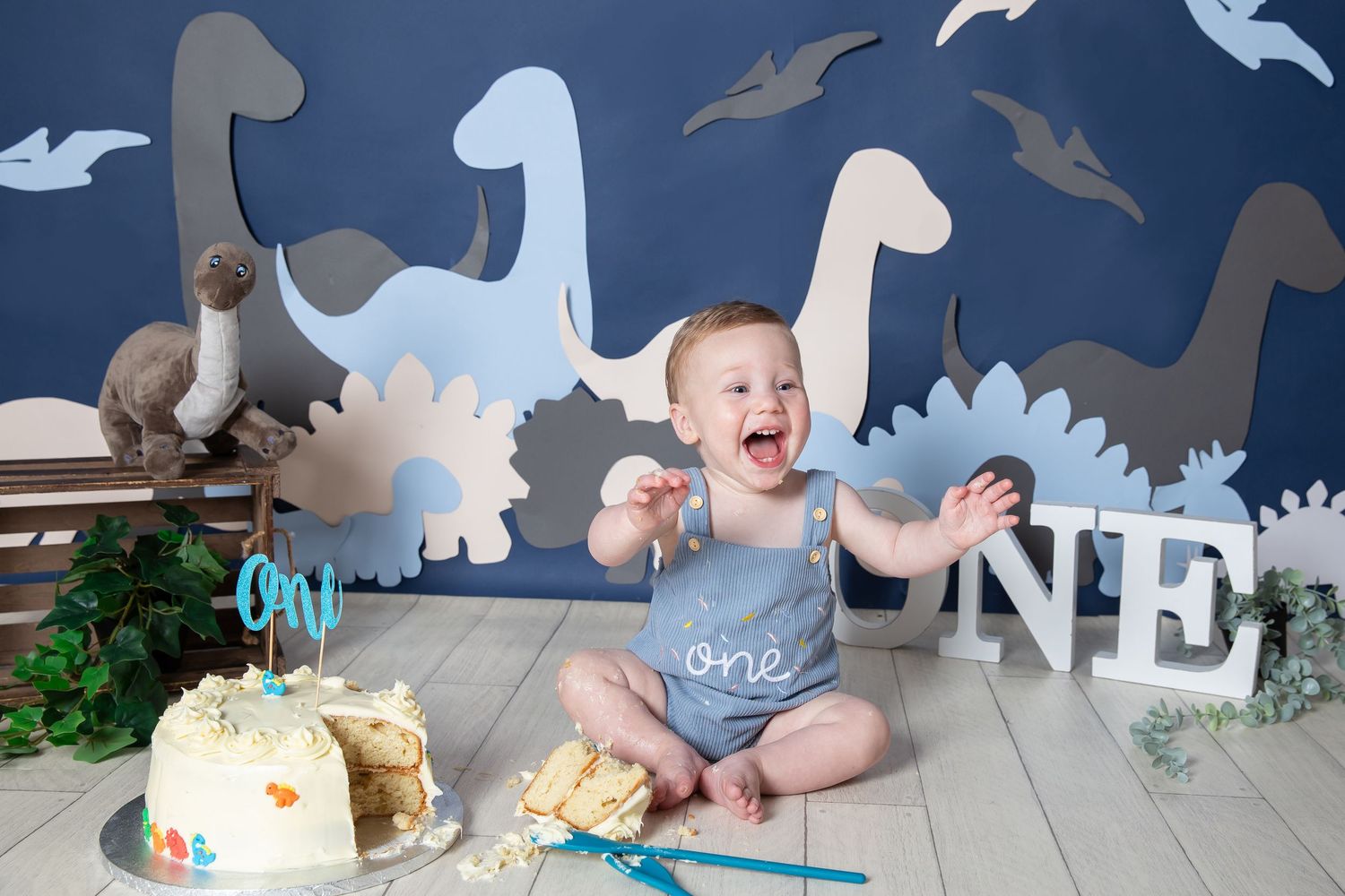 A baby celebrates their first birthday with a dinosaur-themed cake smash photoshoot featuring paper decorations and props.