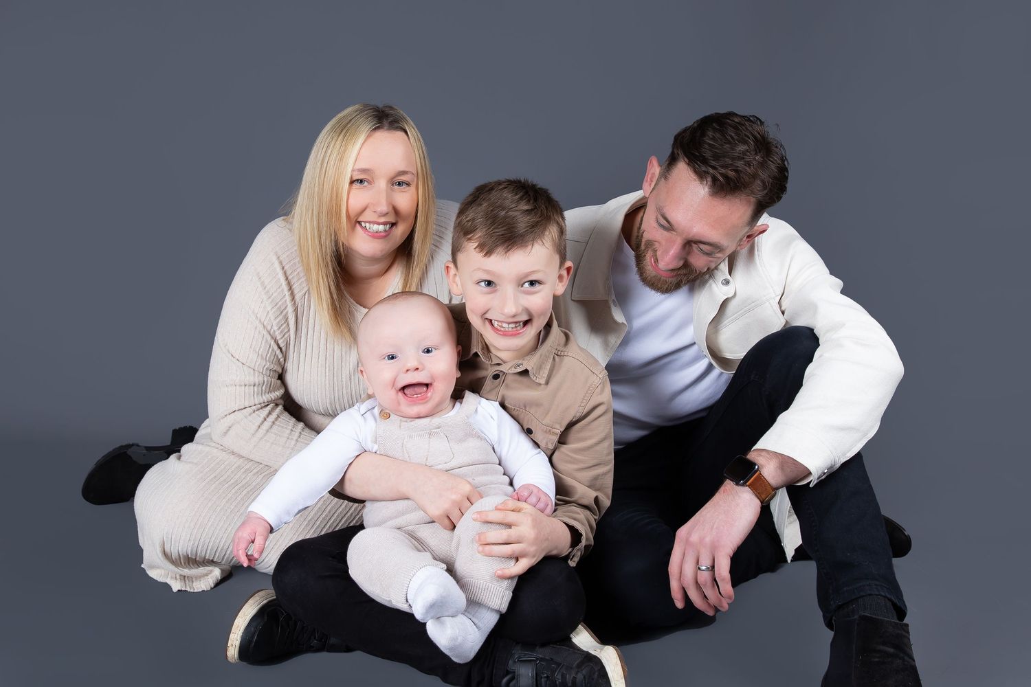 Happy family with young children wearing cream colored clothing posing together against a gray studio backdrop.