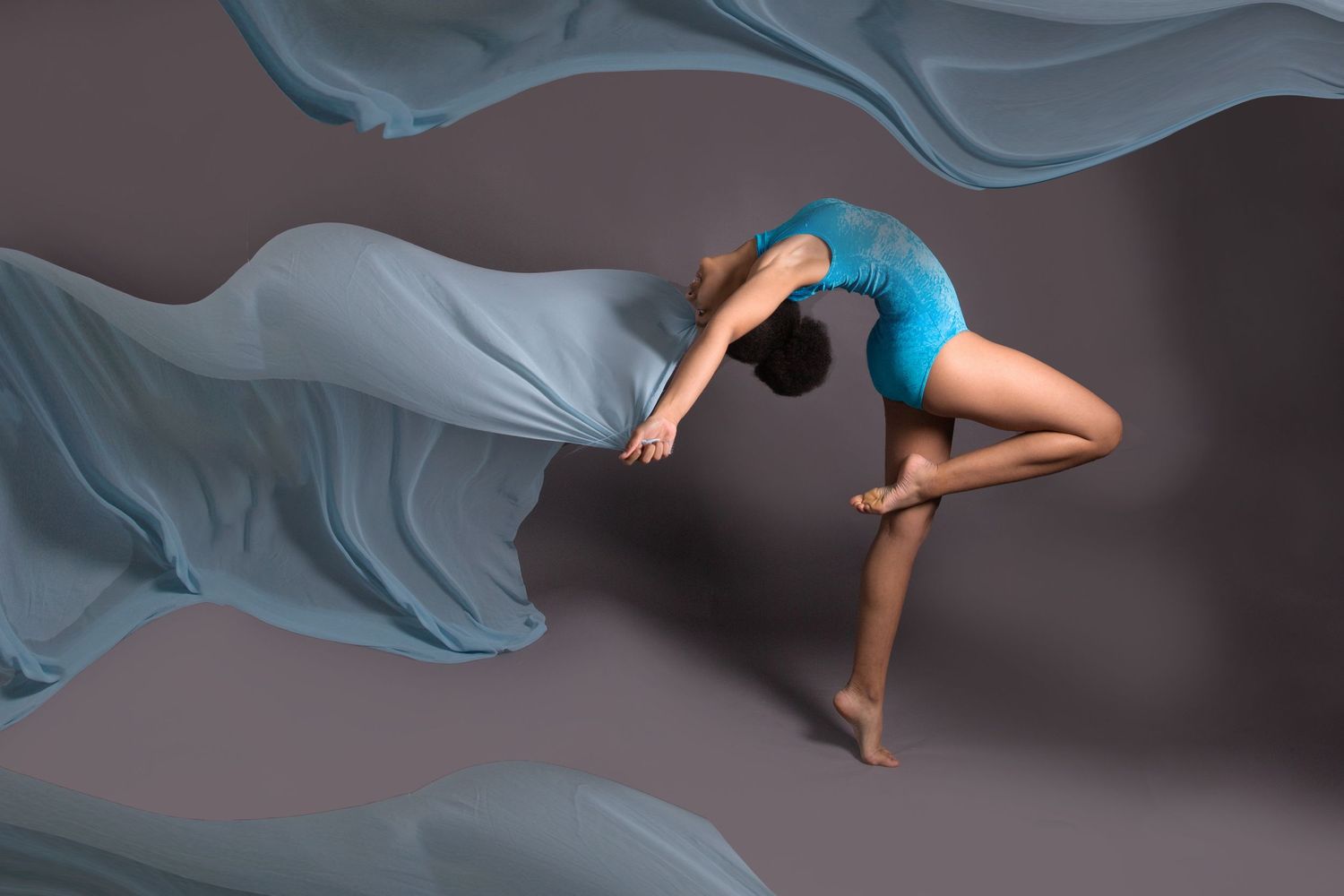 Flowing blue fabric creates artistic movement around a graceful dancer in a dark studio setting.