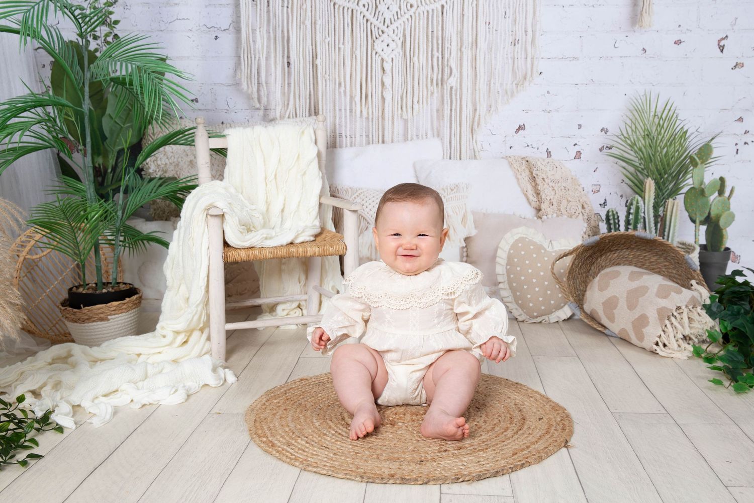 A baby in light pink clothing sits on a round woven rug surrounded by potted plants and rustic decor.