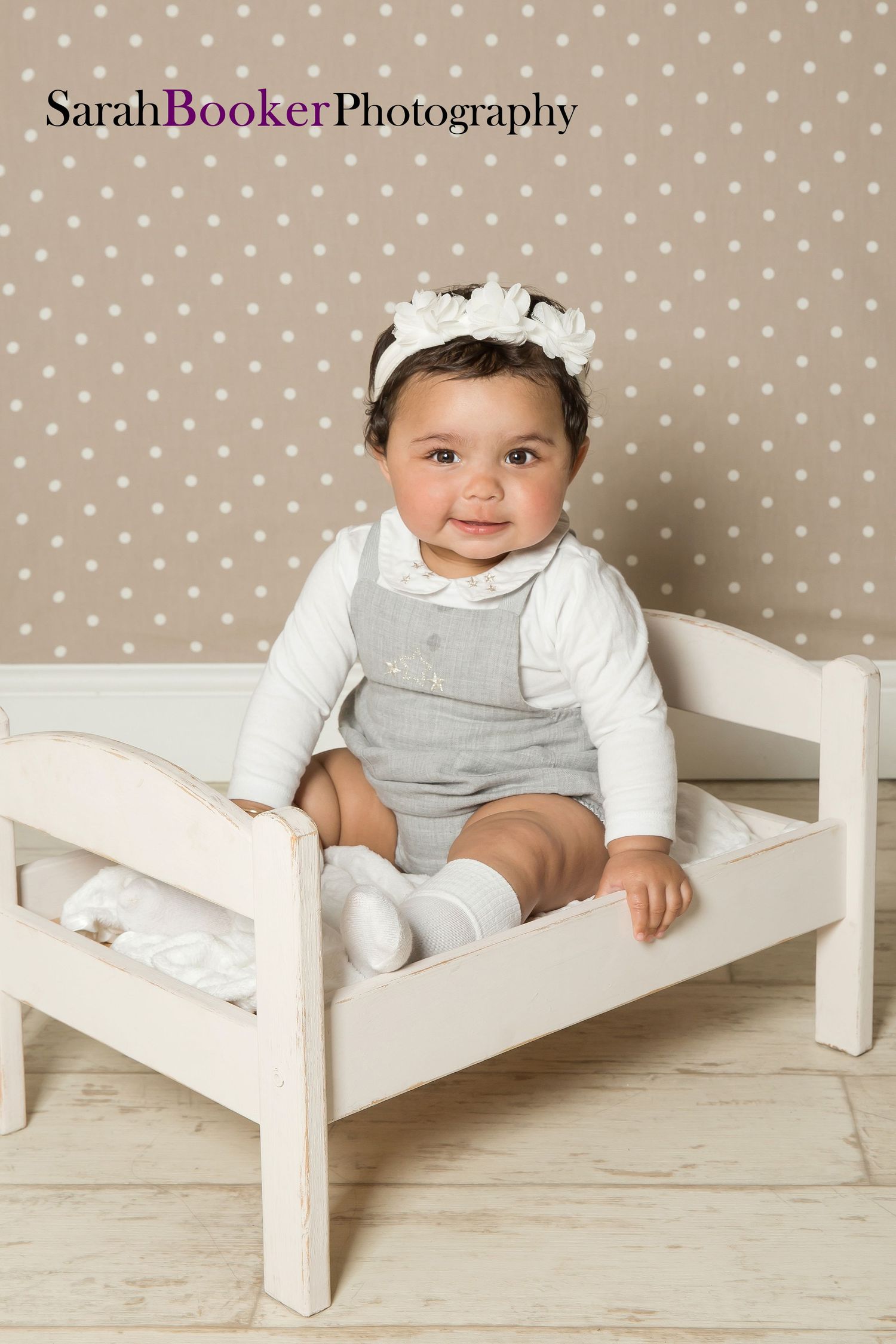 Baby wearing white shirt and gray romper sits on small white wooden bed against polka dot backdrop in studio portrait.