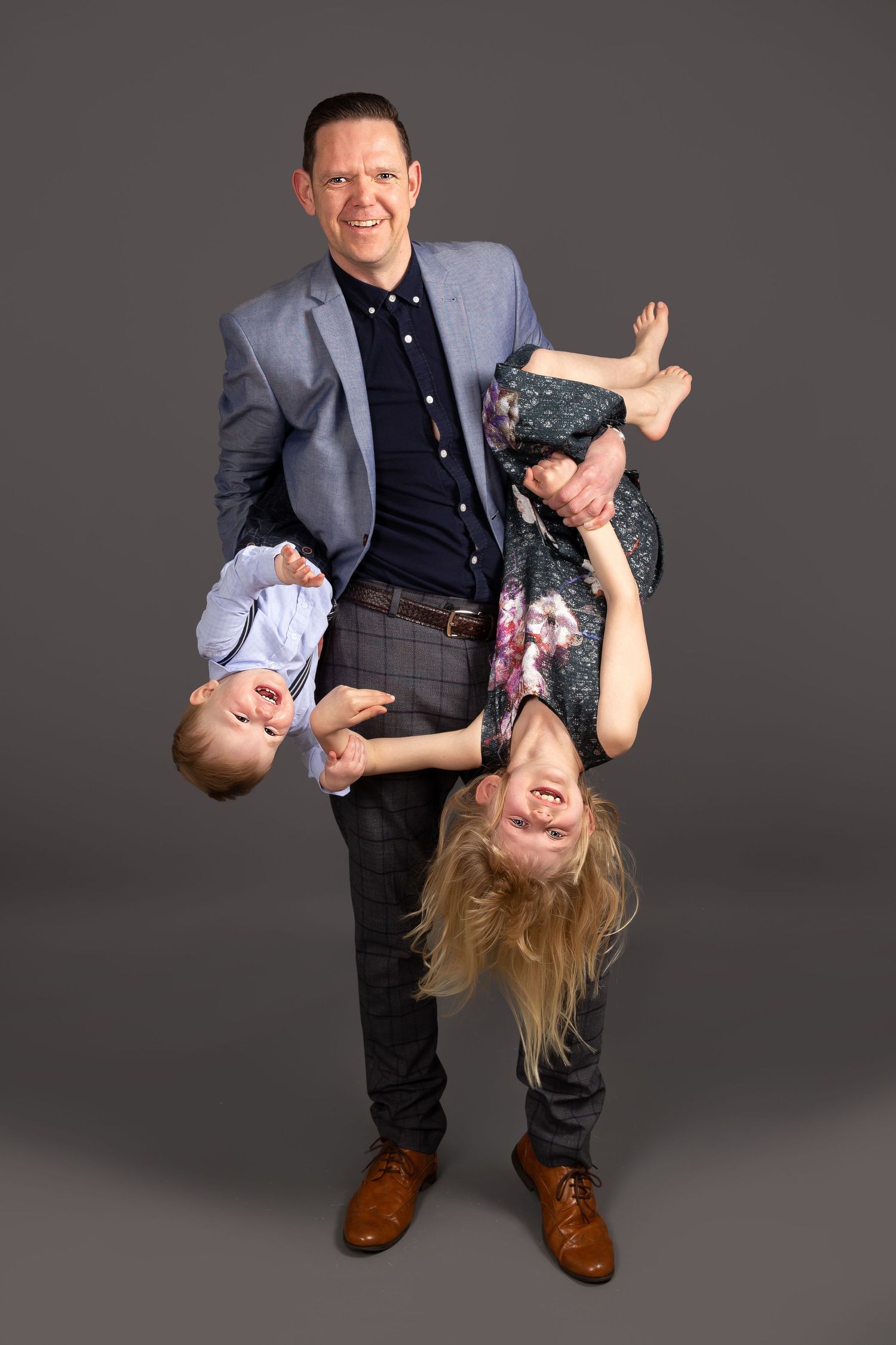 A playful family portrait showing children being held upside down against a dark gray studio backdrop.