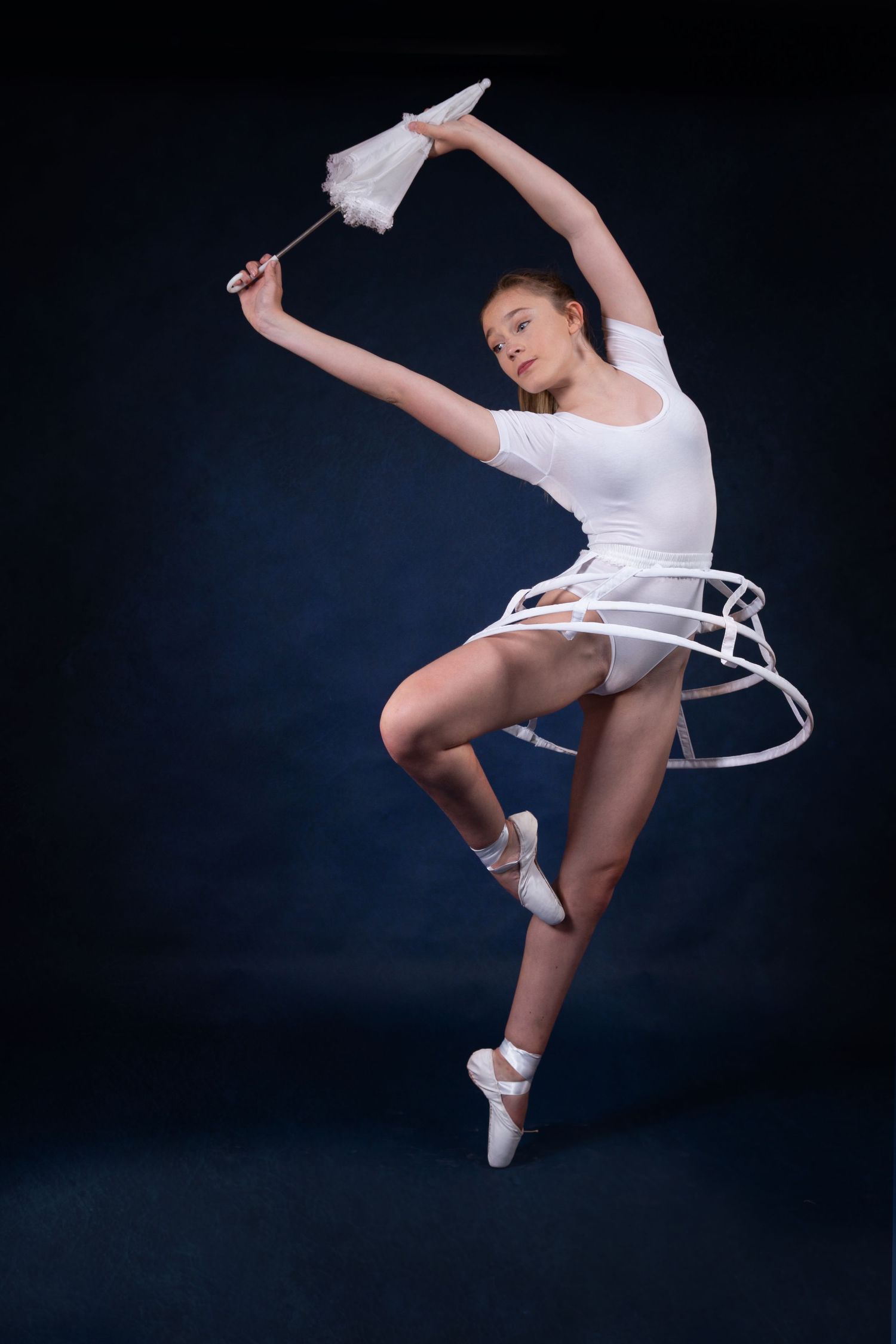 Ballet dancer in white performs graceful leap pose against dark background with artistic hoop element.