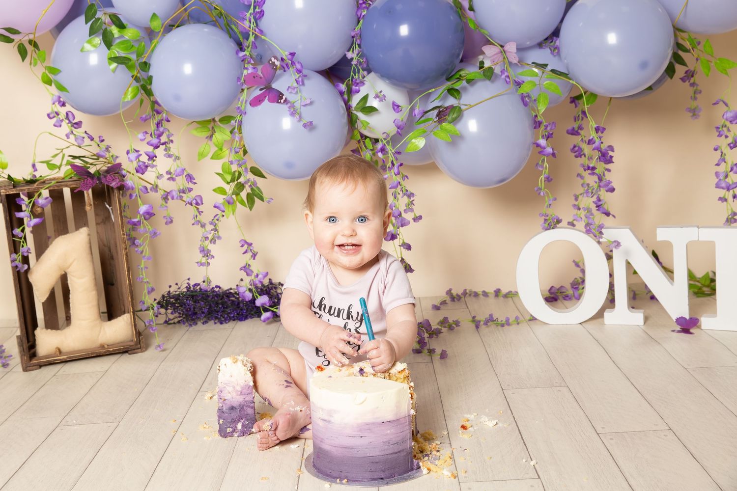 Purple and gray balloon garland decorates a baby's first birthday party setting with floral accents and soft lighting.