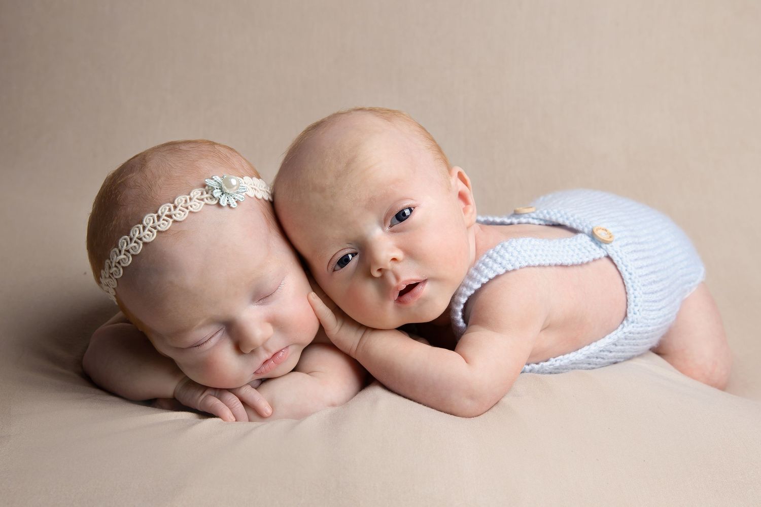 Two sleeping babies in coordinating outfits cuddling together on beige backdrop during portrait session.