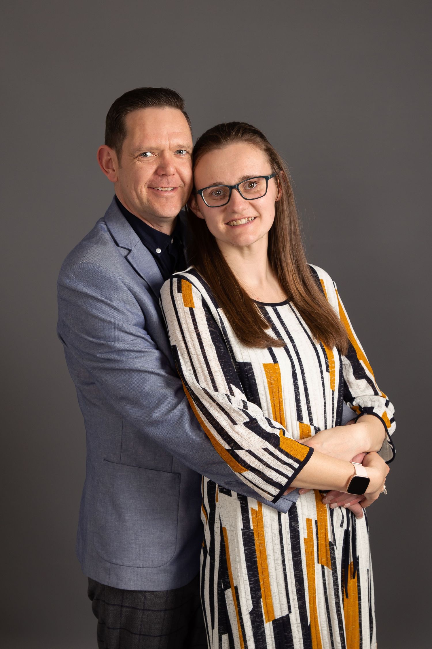 A couple posing for a portrait, with the man in a gray suit and the woman in a striped dress against a gray background.