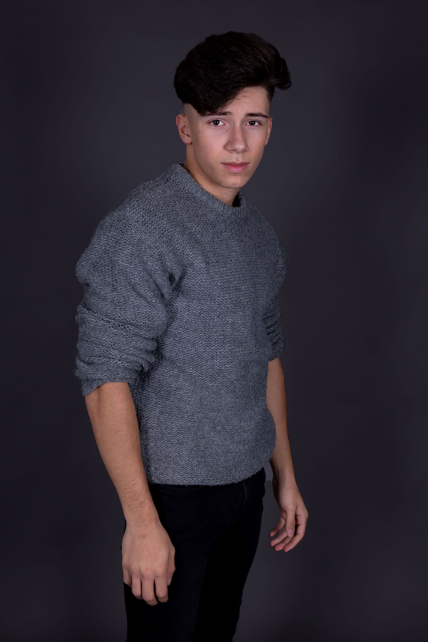 Studio portrait of a young person in a casual grey sweater and dark pants against a black background.