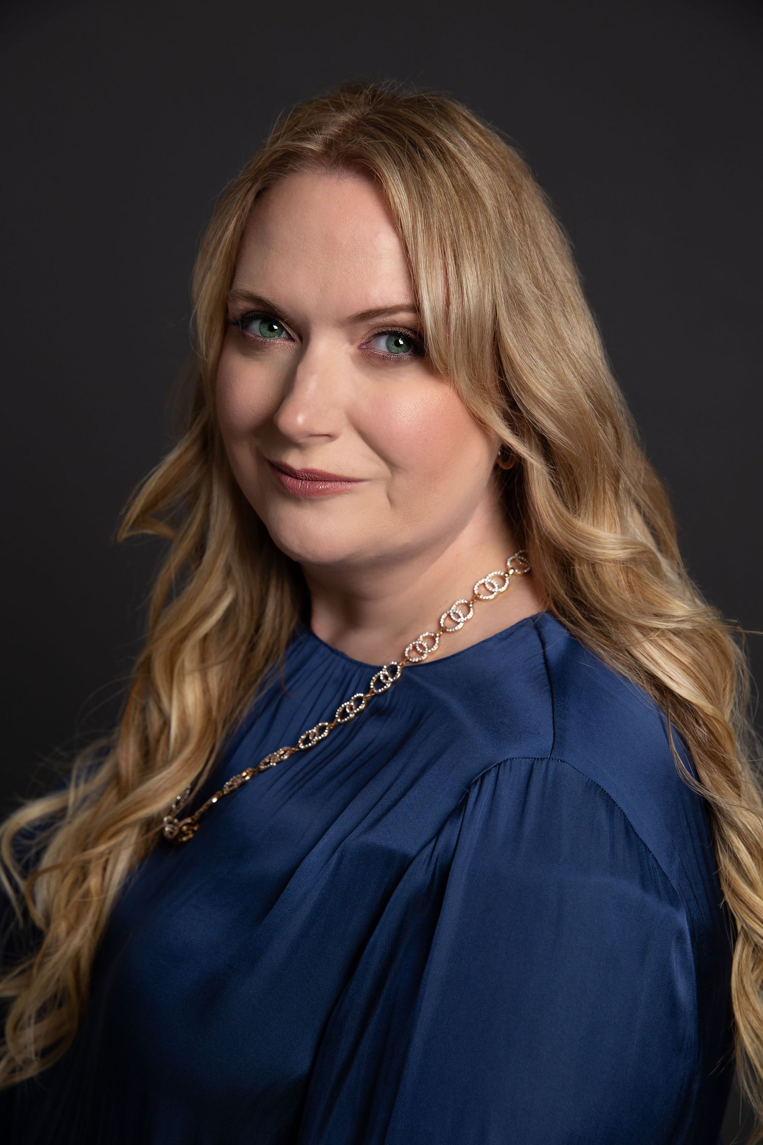 Professional headshot featuring a subtle smile and blonde wavy hair styled with a navy blue blouse against dark background.