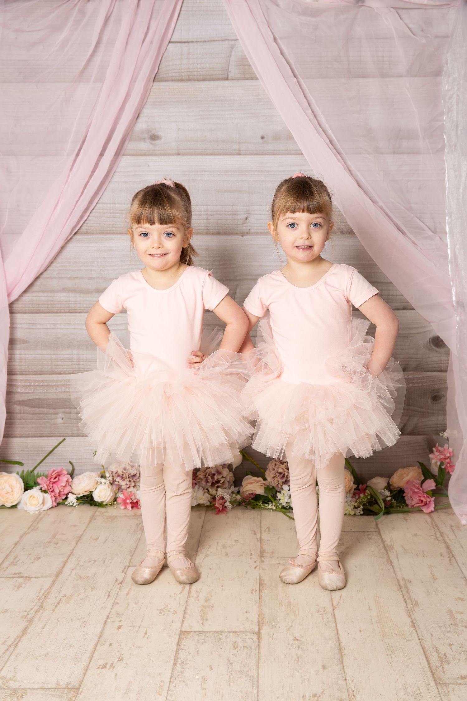 Two young ballet dancers in pink tutus and leotards pose in front of a draped backdrop with flowers.
