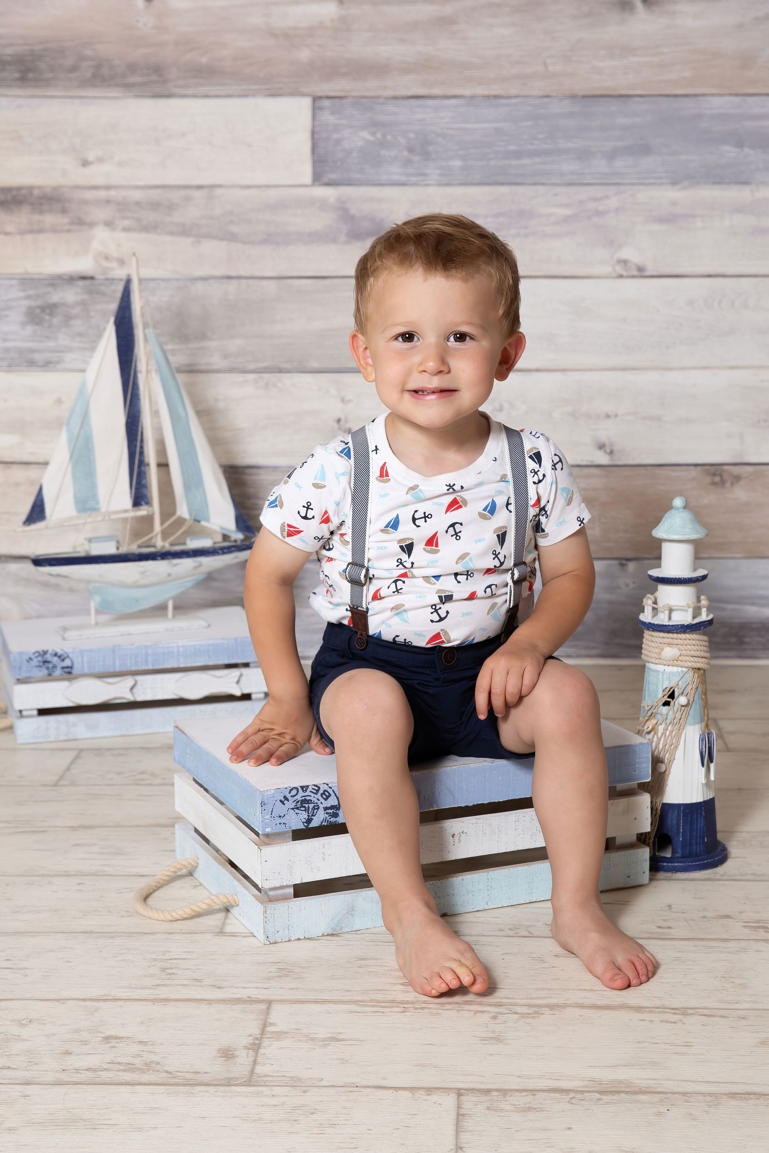 Young child sits on stacked books with nautical decor including toy sailboat and lighthouse against rustic wood wall.
