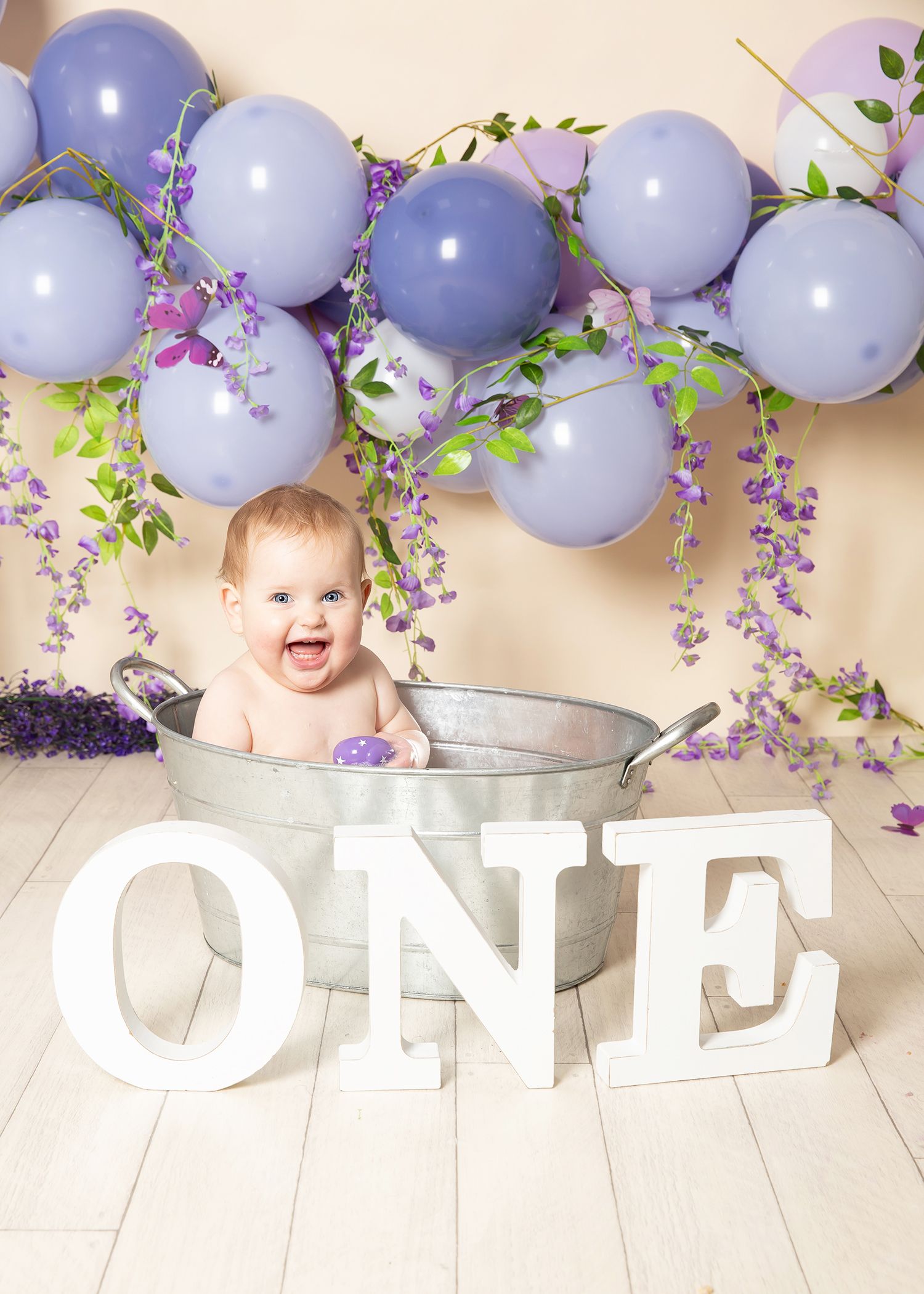Festive purple and lavender balloon garland decorates a wall with floral accents and baby-themed decor.