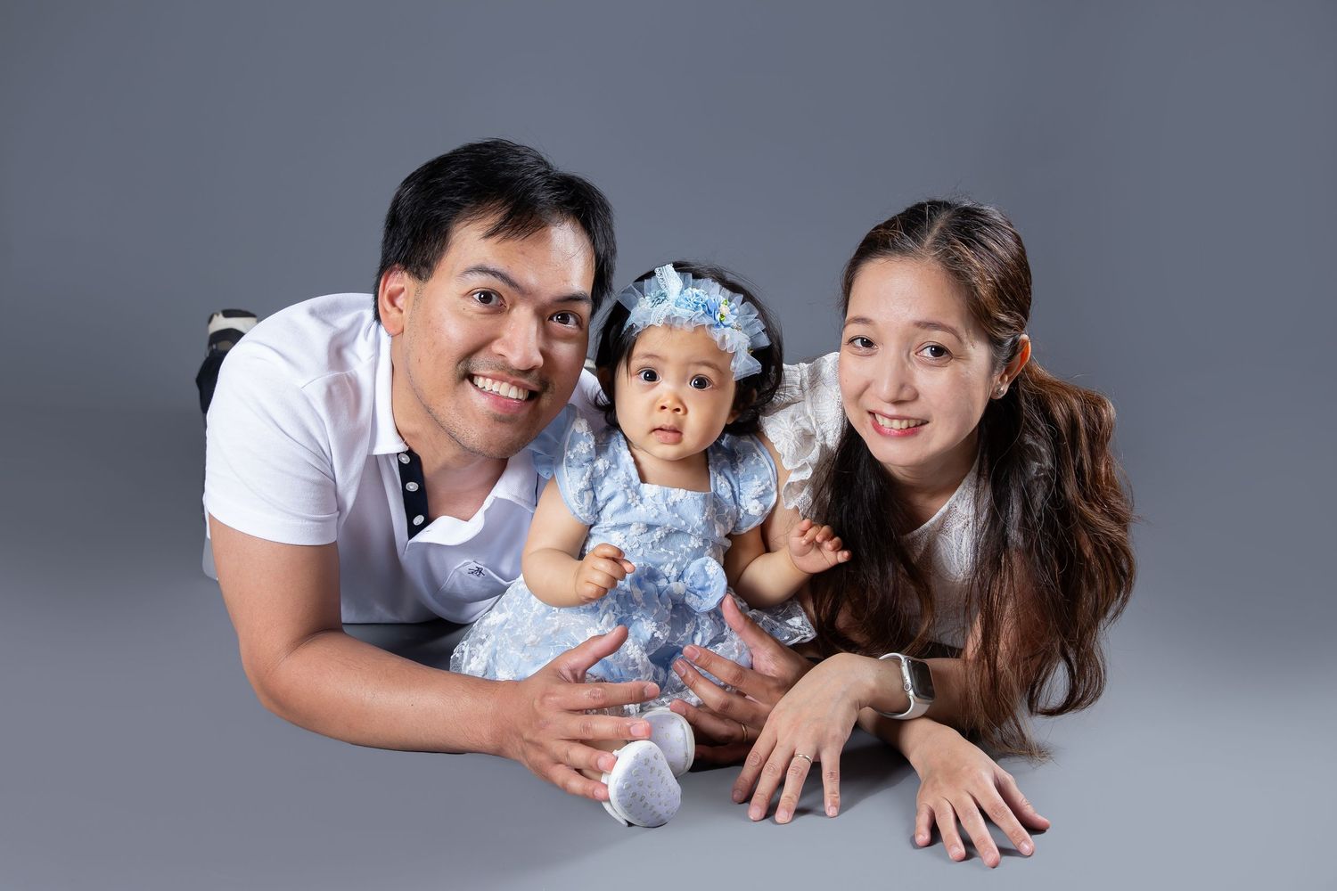 Family in white clothing lying together on grey background during professional photo session.