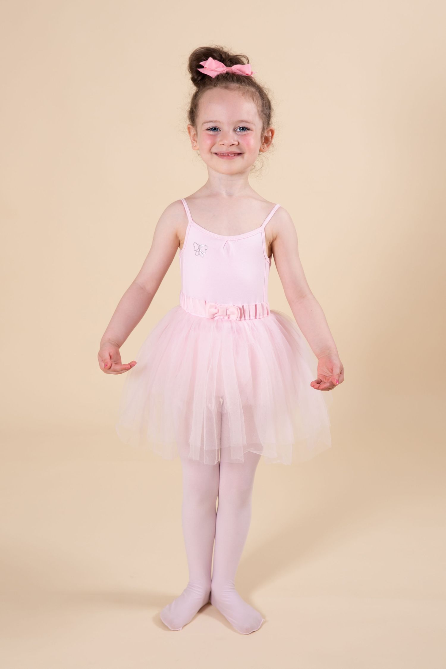 Young ballet dancer in pink tutu and tights stands in first position against beige background.